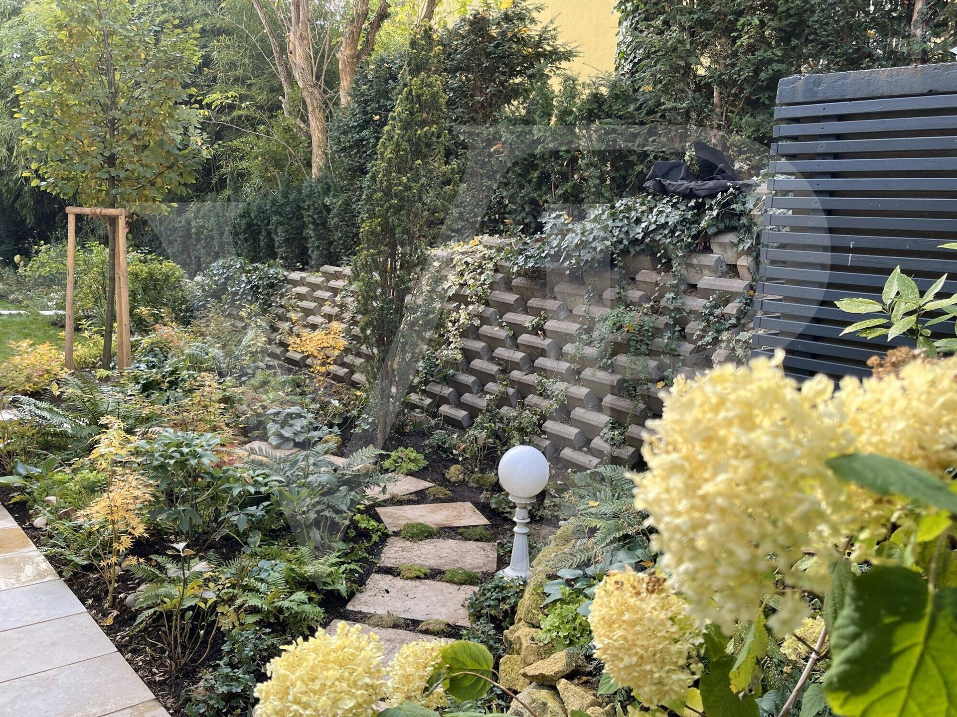 Terrassierter Garten mit Steinmauern, üppiger Bepflanzung und einem dekorativen Lichtobjekt.