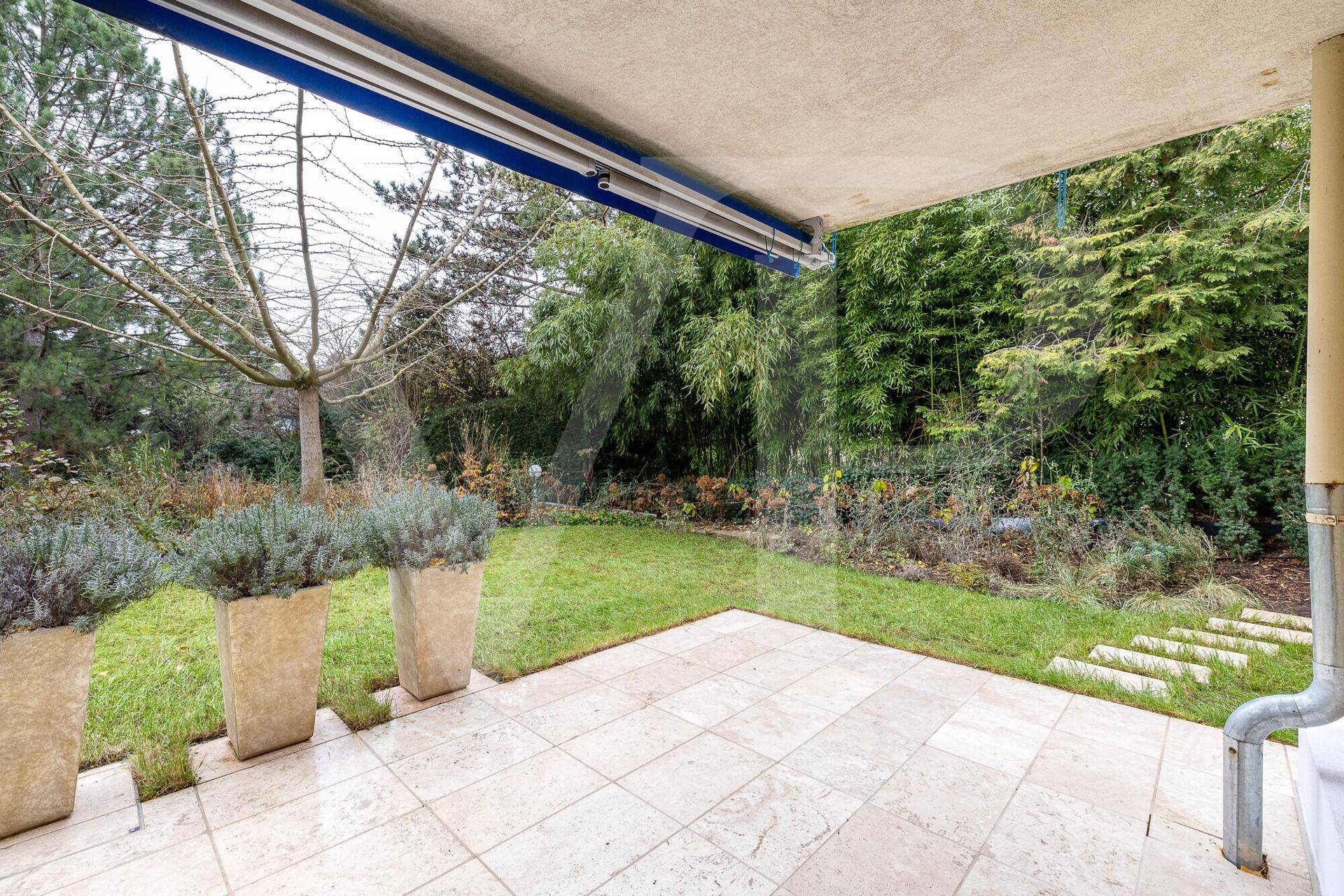 Überdachte Terrasse mit Fliesenboden und Blick auf den grünen Garten mit Bäumen.
