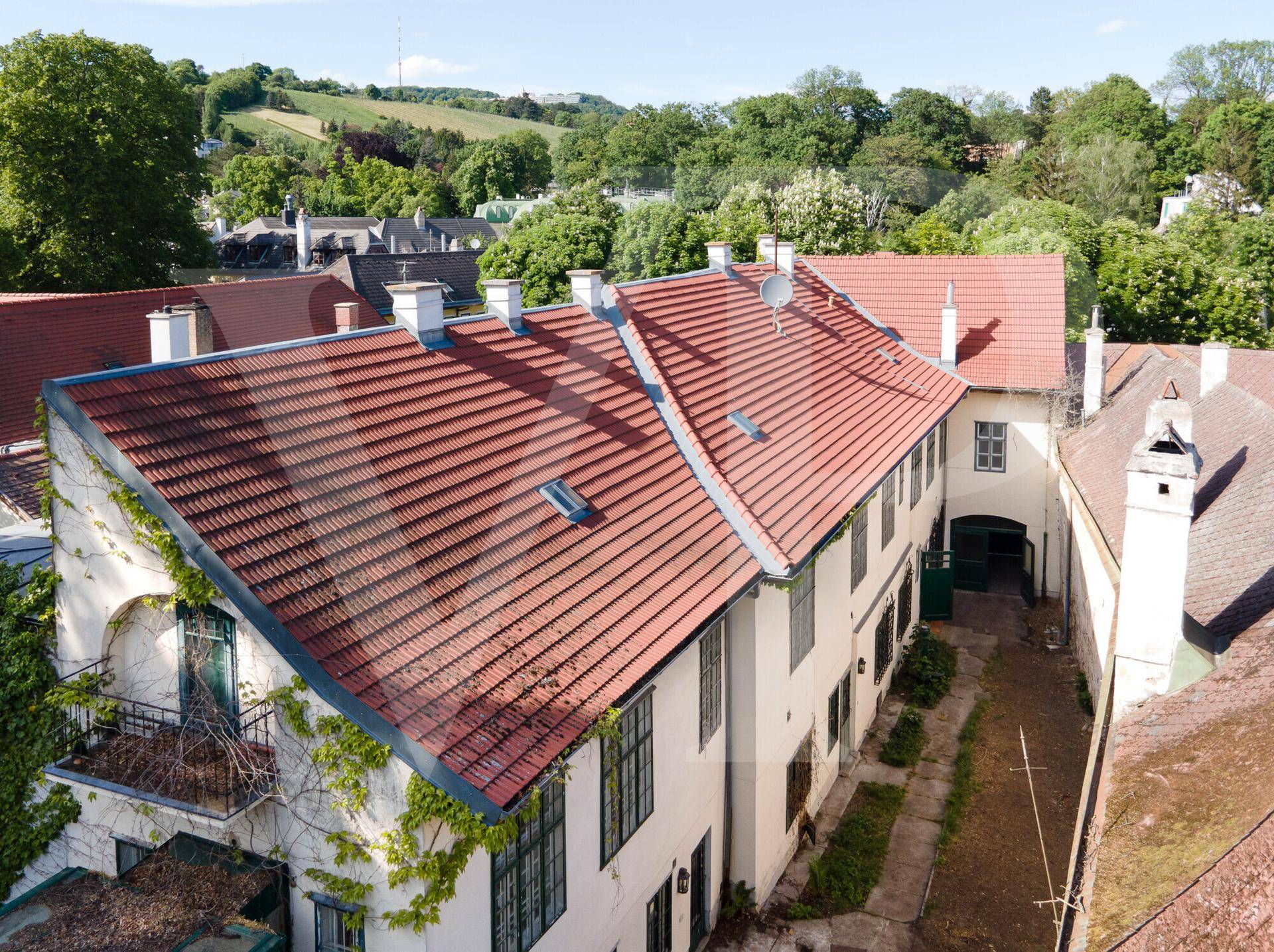 Historisches Gebäude mit rotem Ziegeldach und Schornsteinen, umgeben von üppiger grüner Landschaft.