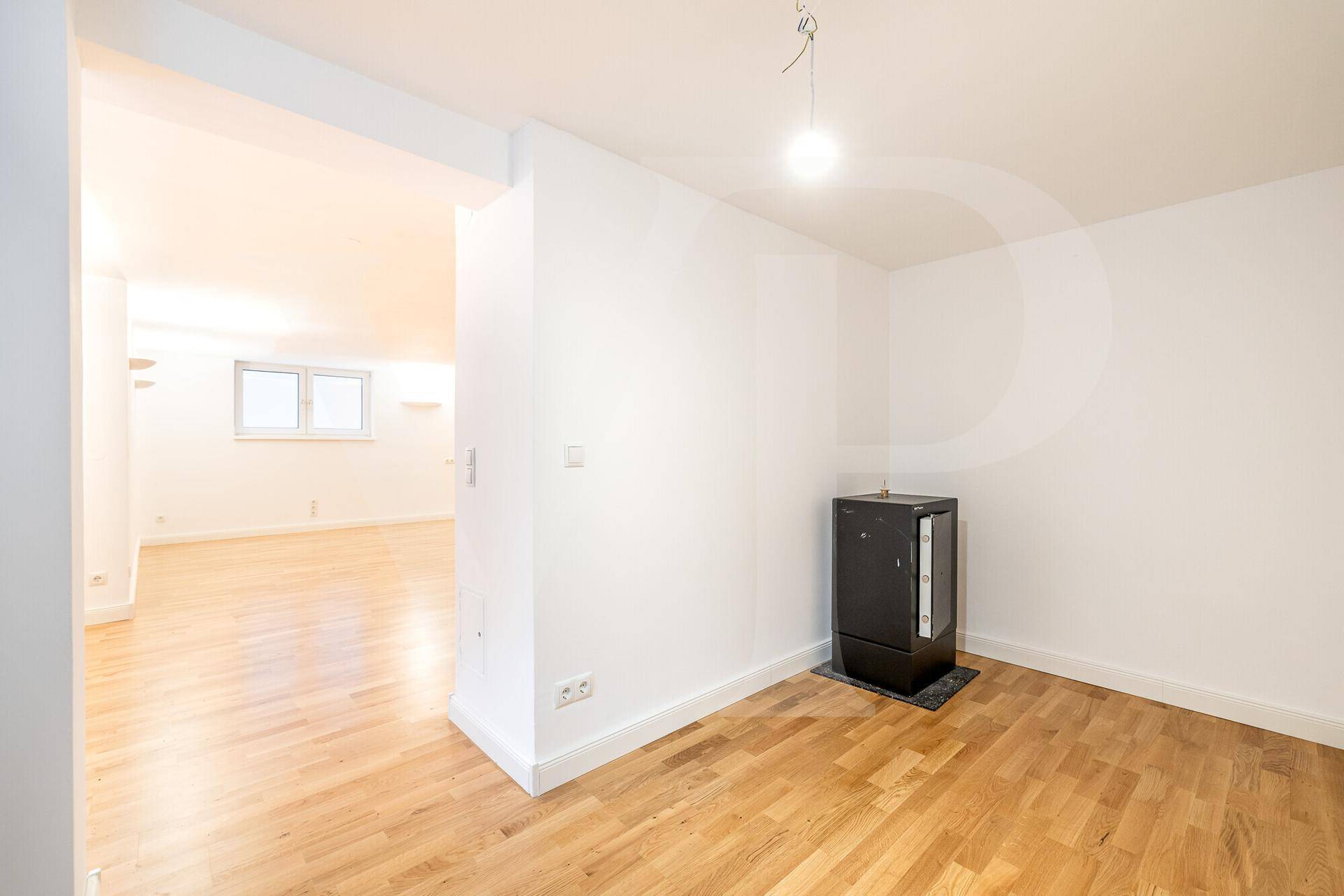 Helles Zimmer mit Holzboden und einem modernen Ofen, offen zu einem weiteren Raum mit Fenstern.