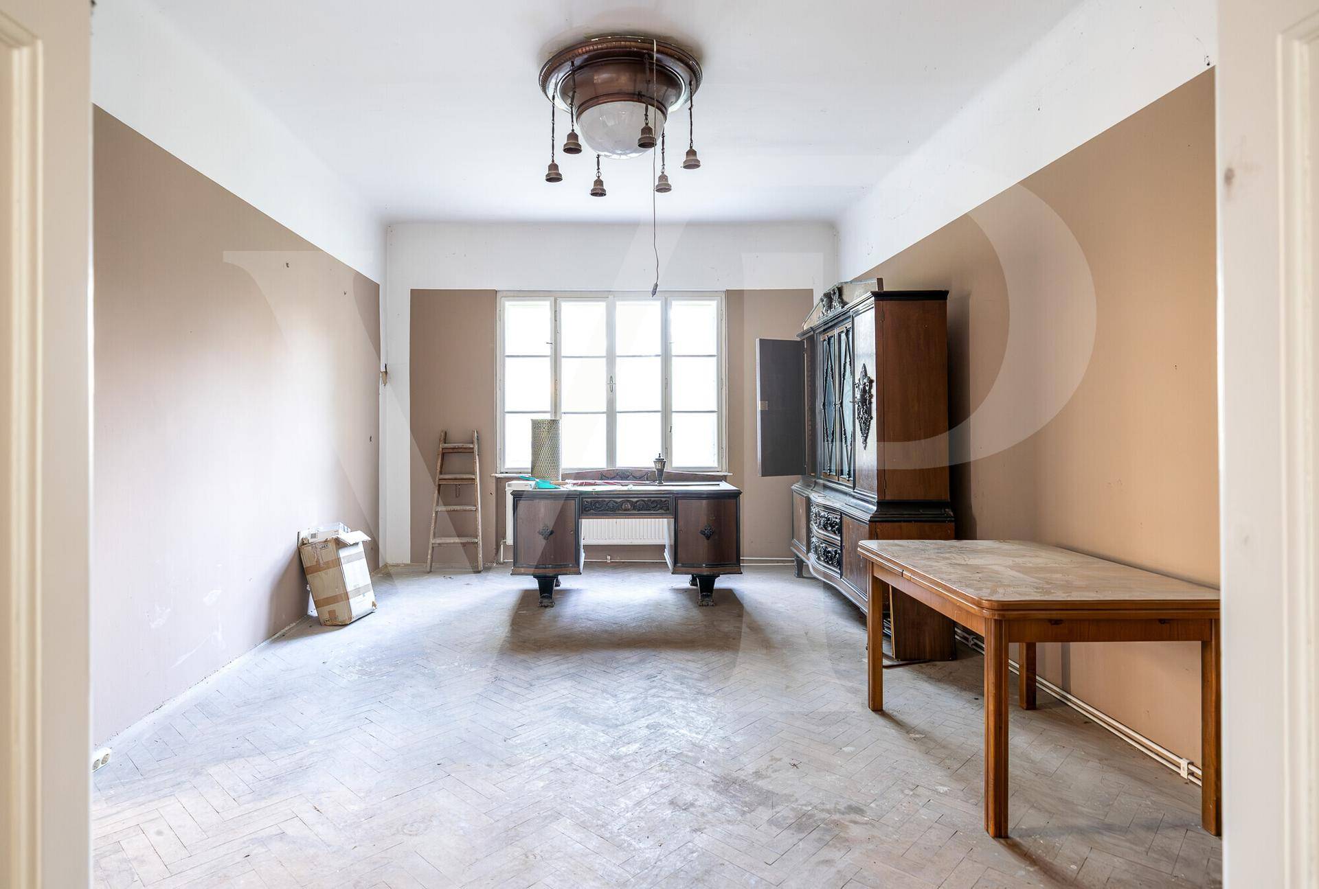 Zimmer mit abgenutztem Fischgrätparkett, zweifarbigen Wänden, antikem Schreibtisch und Schrank.
