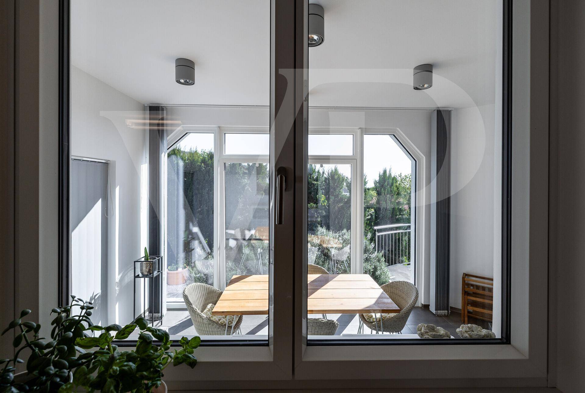 Blick durch ein Fenster auf einen hellen Essbereich mit Holztisch und Korbstühlen.