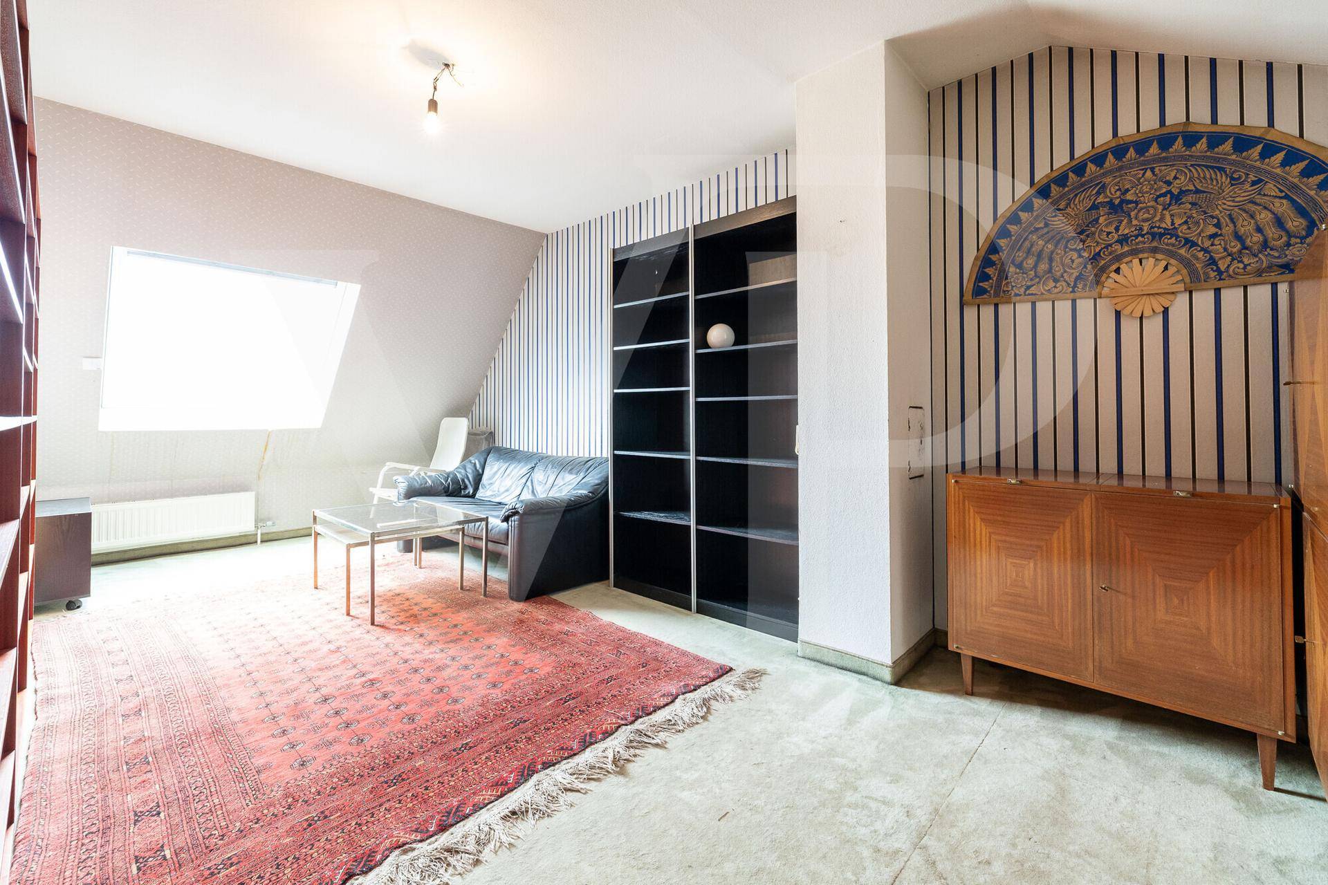 Geräumiges Dachzimmer mit Dachfenster, Teppich und schwarzem Sofa, bietet vielseitige Nutzungsmöglichkeiten.