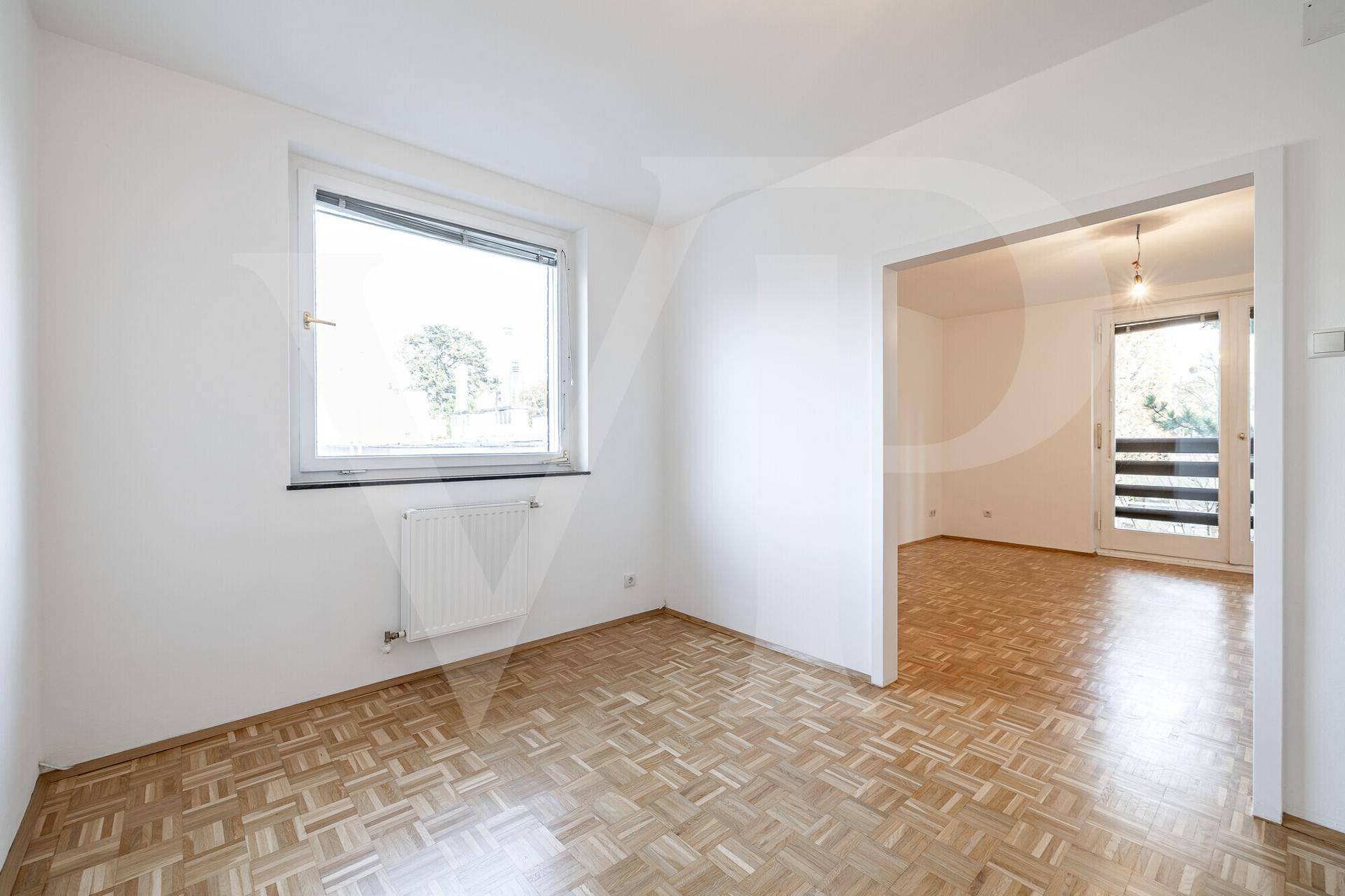 Leeres Zimmer mit Parkettboden, großen Fenstern und Zugang zu einem weiteren Wohnbereich.