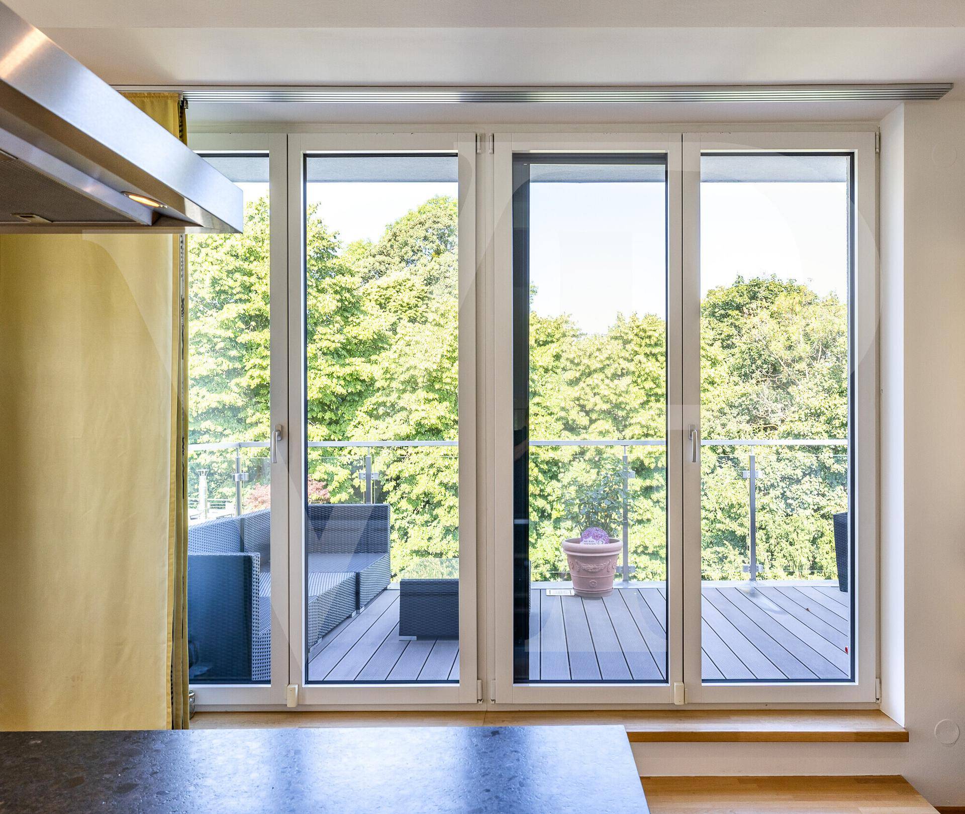 Blick von der Küche durch große Fenster auf eine möblierte Terrasse mit grüner Umgebung.