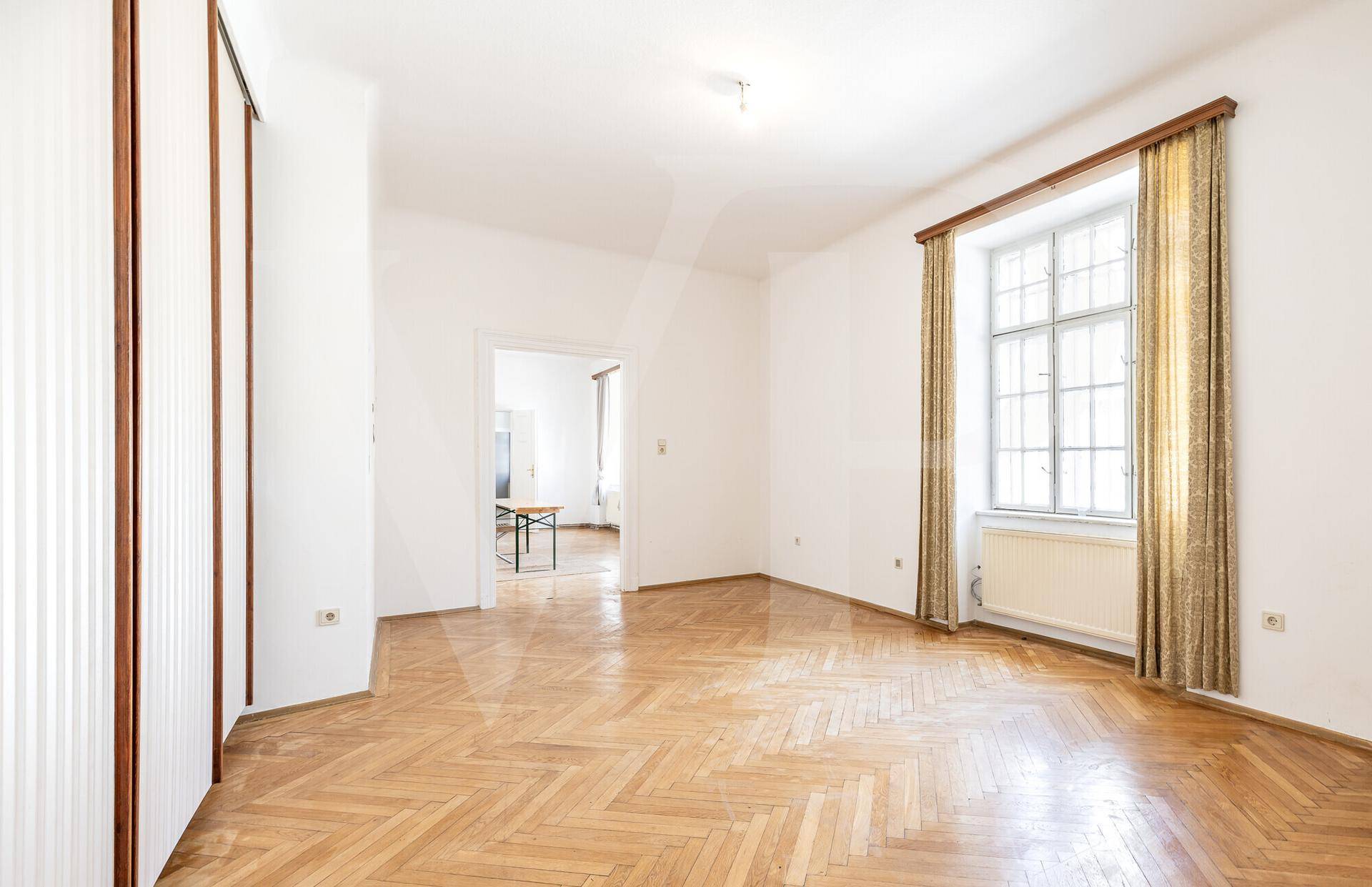 Helles, leeres Zimmer mit Fischgrätparkett, hohen Decken und großen Fenstern.