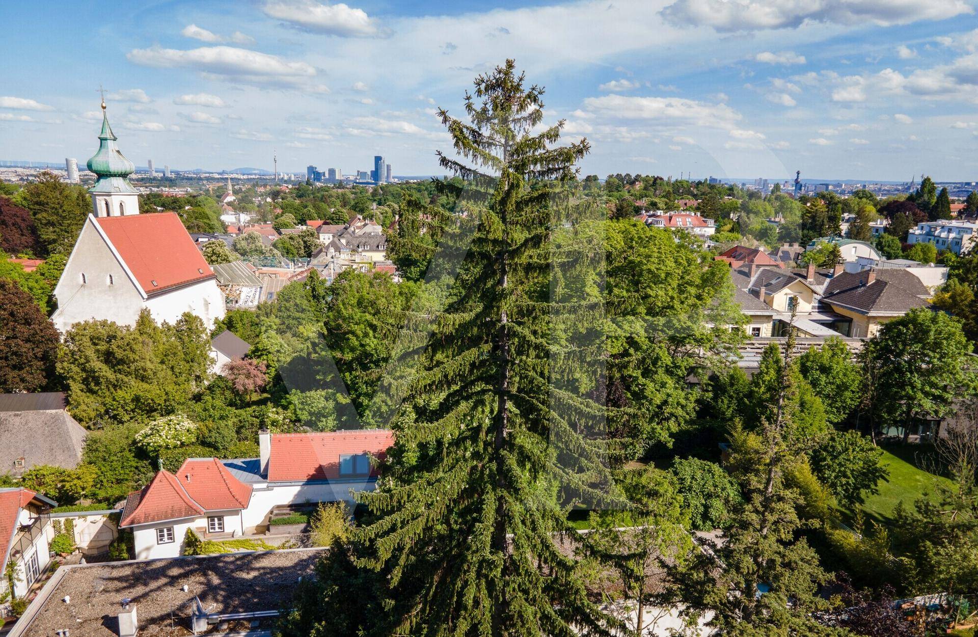 Weitläufige Aussicht über eine Stadt mit Kirche, hohen Bäumen und einer fernen Skyline.