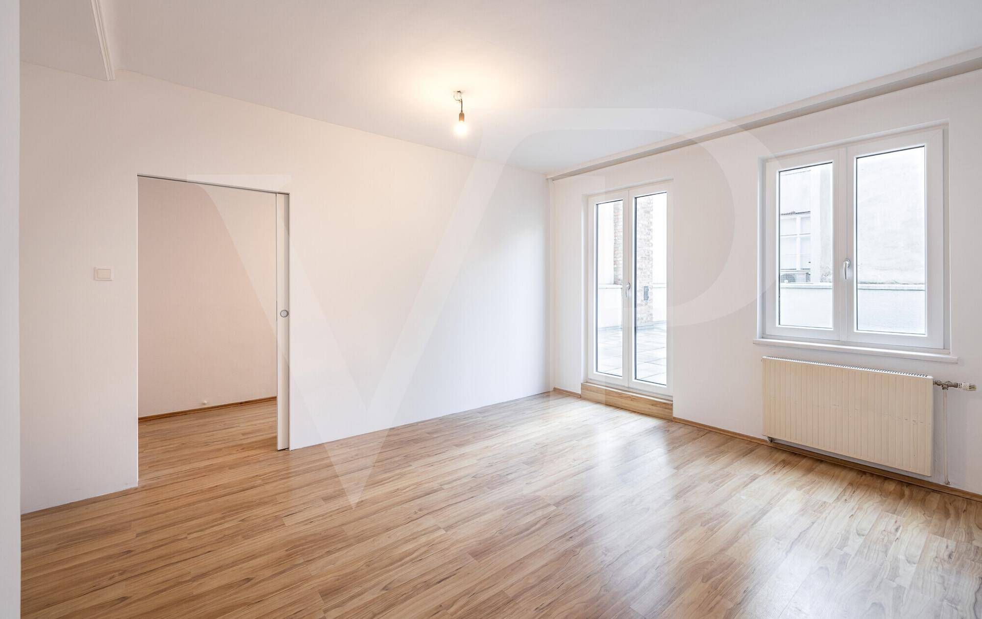 Heller, leerer Wohnraum mit Laminatboden, großen Fenstern und Zugang zu einem Balkon oder einer Terrasse.
