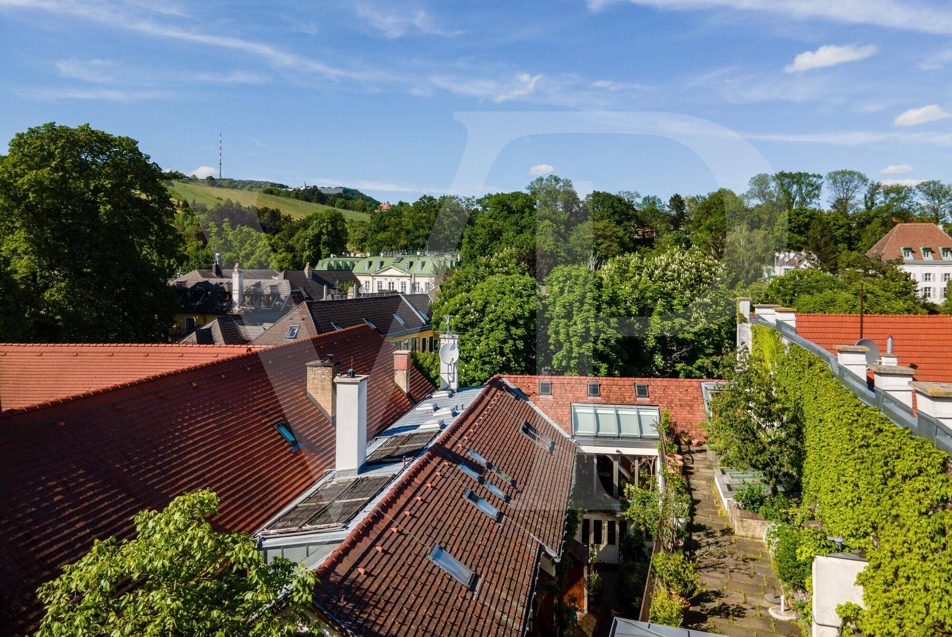 Blick über rote Ziegeldächer auf eine grüne Hügellandschaft unter blauem Himmel.