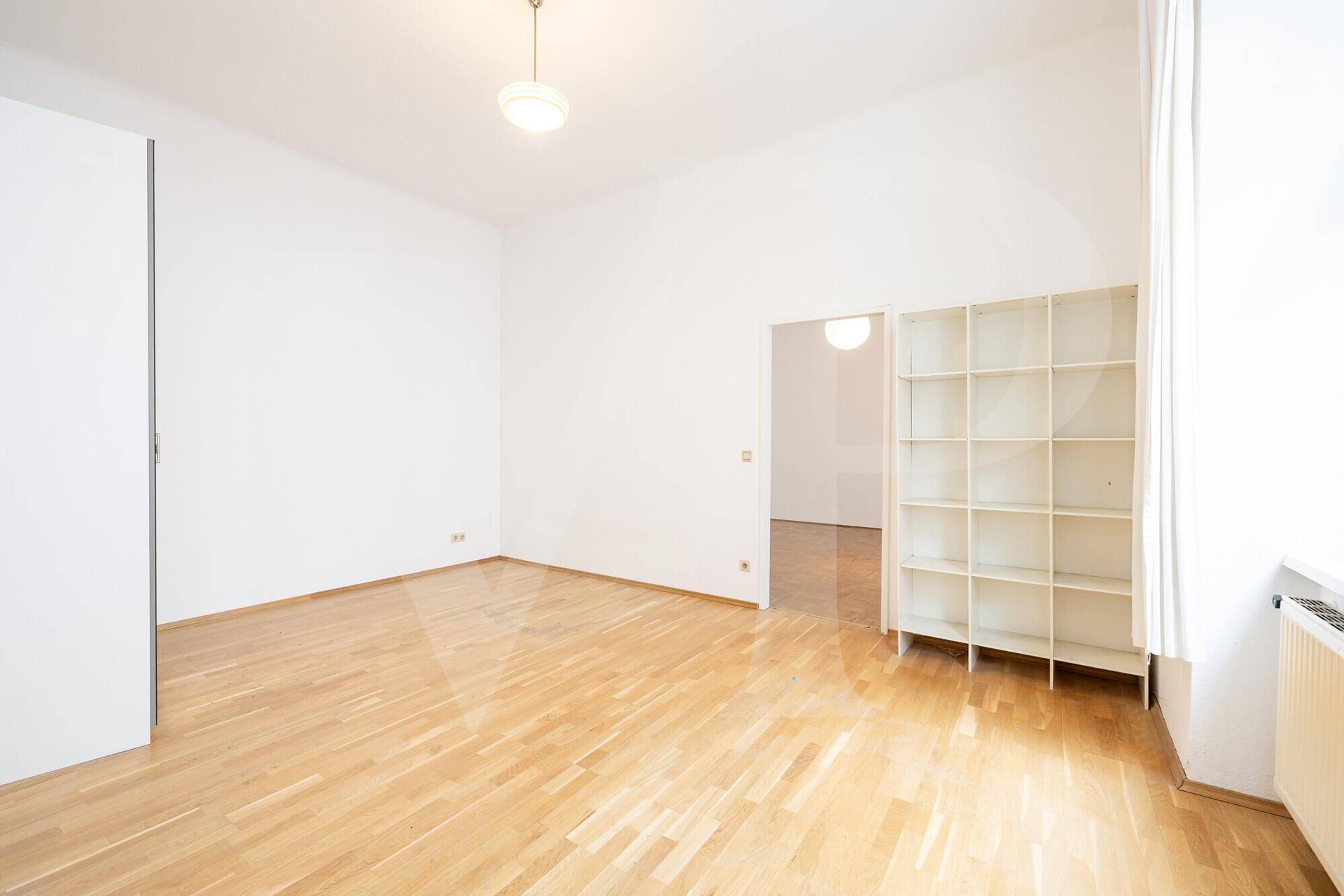 Helles Zimmer mit Parkettboden, einem Schrank und einem offenen Durchgang zu einem Nebenraum.