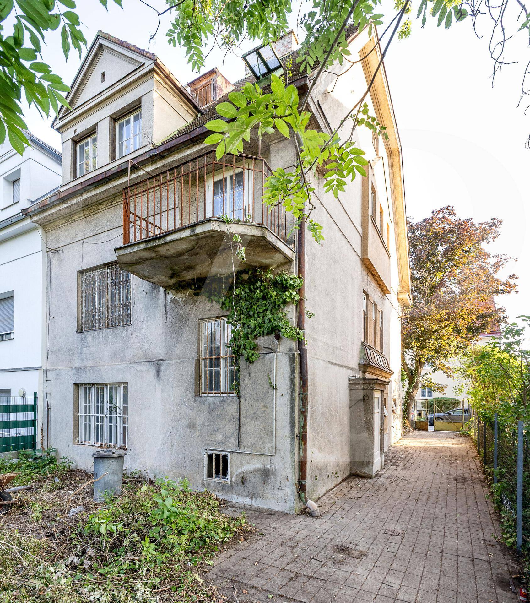 Seitenansicht des Gebäudes in unrenoviertem Zustand mit verwitterter Fassade und überwuchertem Gartenbereich.