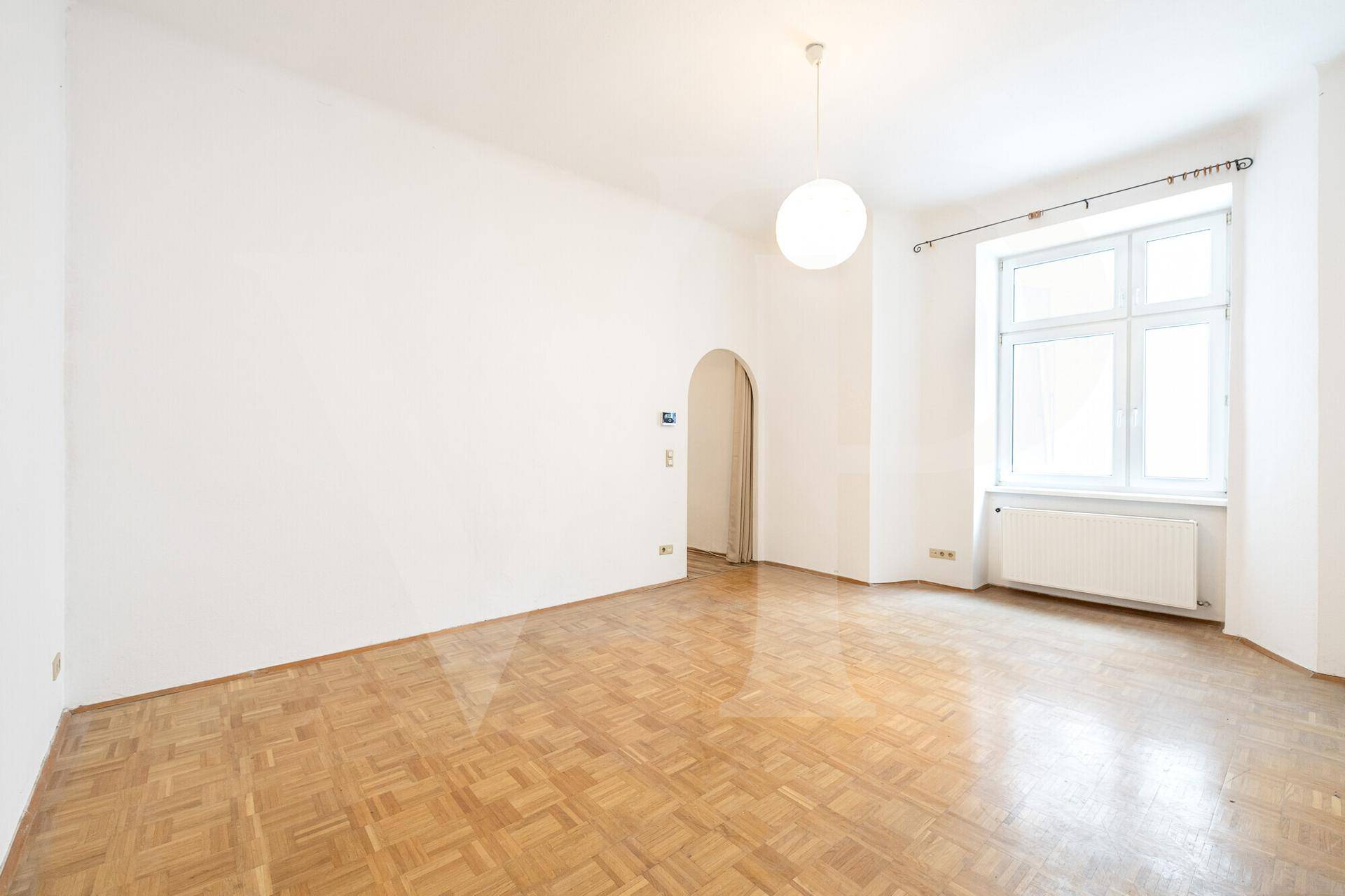 Heller Raum mit Parkettboden, großem Fenster und einem Rundbogen, der zu einem weiteren Bereich führt.