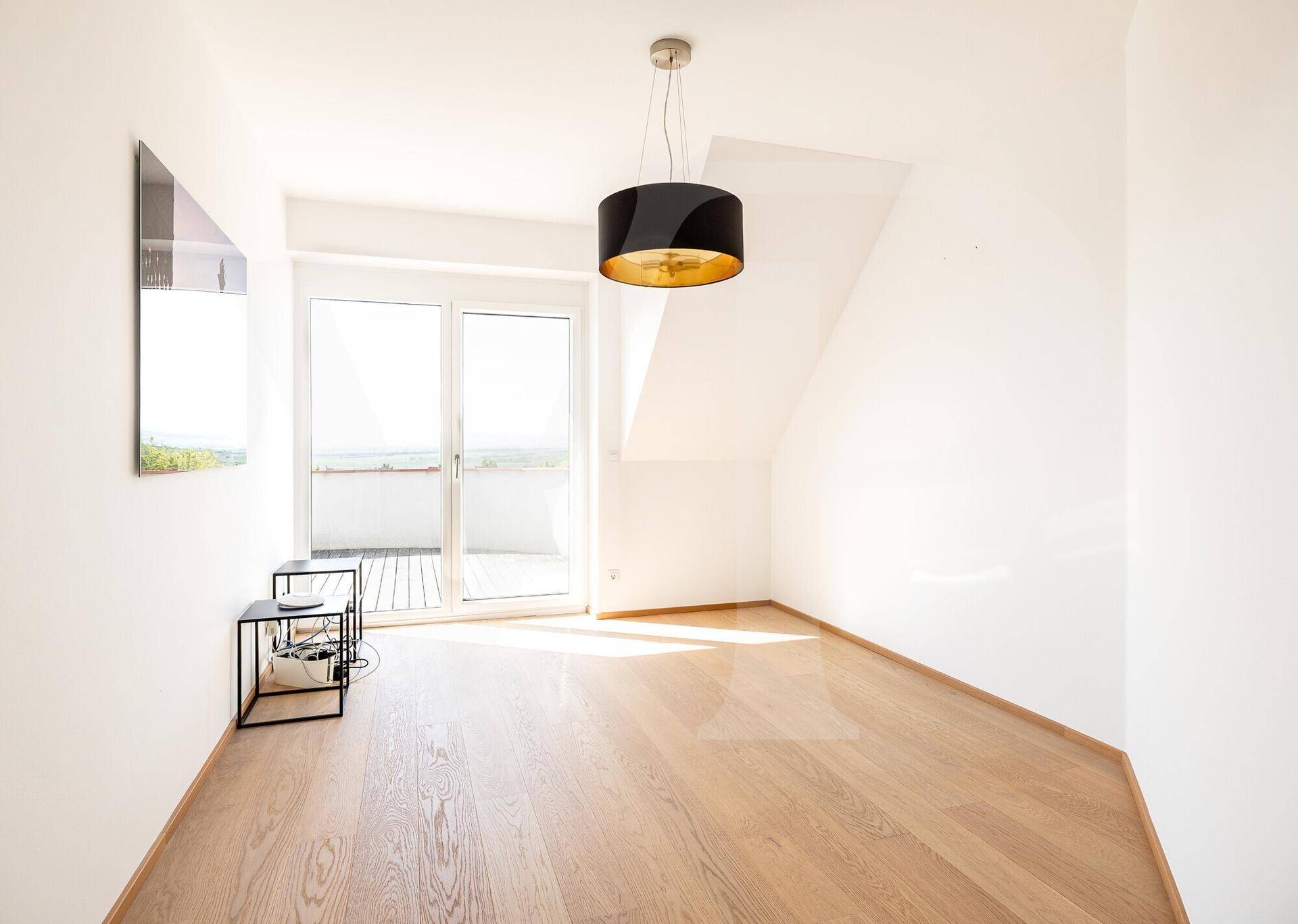 Heller Raum mit Holzboden, Dachschräge und Zugang zu einem kleinen Balkon.