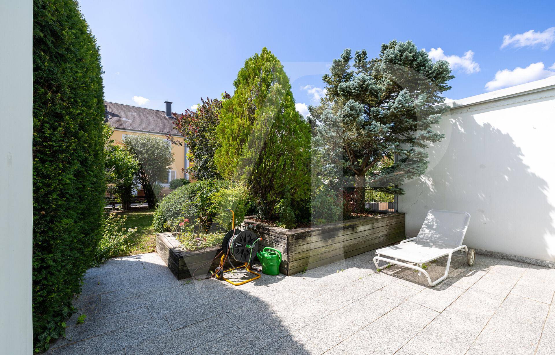 Terrasse mit Steinplatten, erhöhten Blumenbeeten und Liegestuhl, umgeben von Grün.