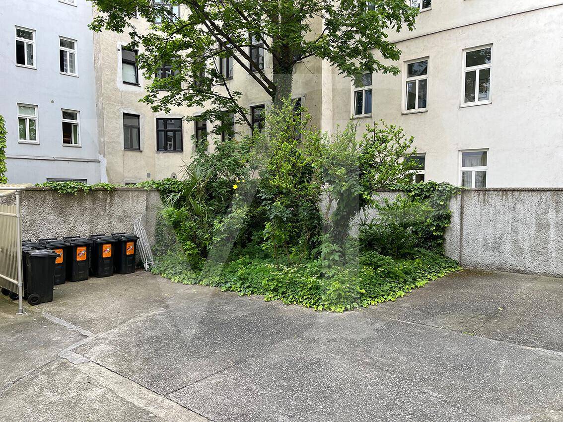 Kleiner begrünter Innenhof mit Bäumen, Sträuchern und Mülltonnen.