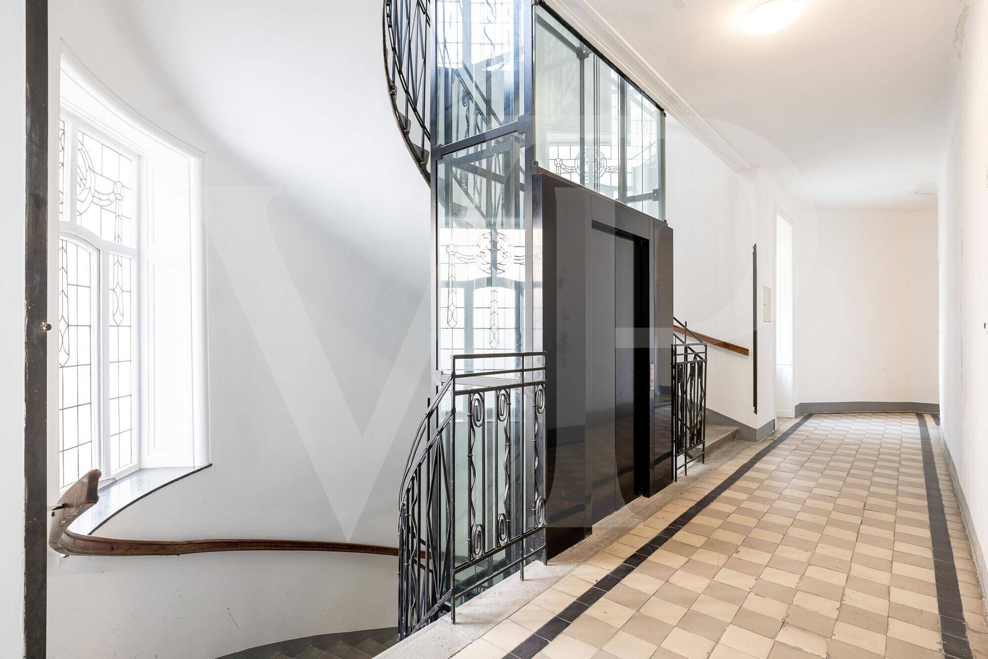 Heller Treppenhausbereich mit elegantem Geländer, Aufzug und gemustertem Fliesenboden.
