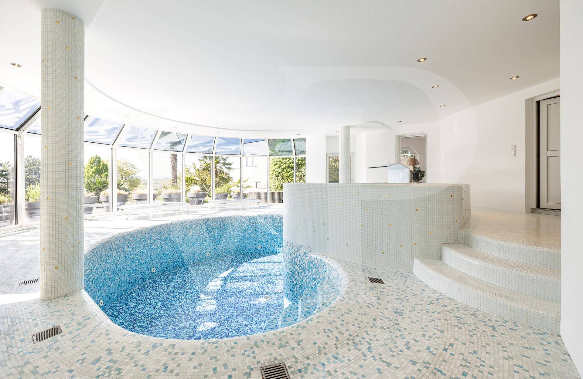 Heller Innenpoolbereich mit Mosaikfliesen, großen Fenstern und Blick ins Grüne.