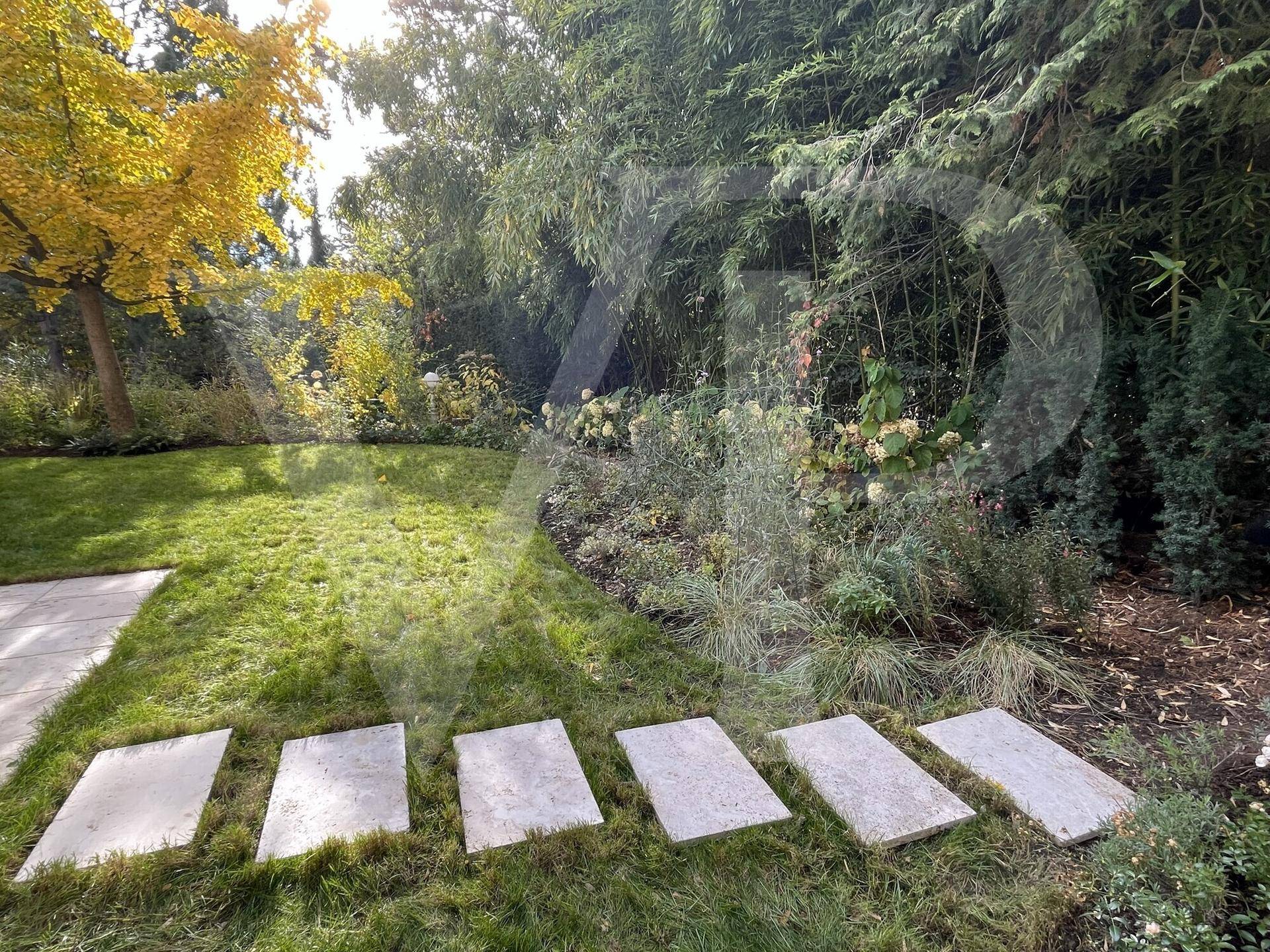 Garten mit Rasenfläche und Trittsteinen, umgeben von üppiger Vegetation und hohen Bäumen.