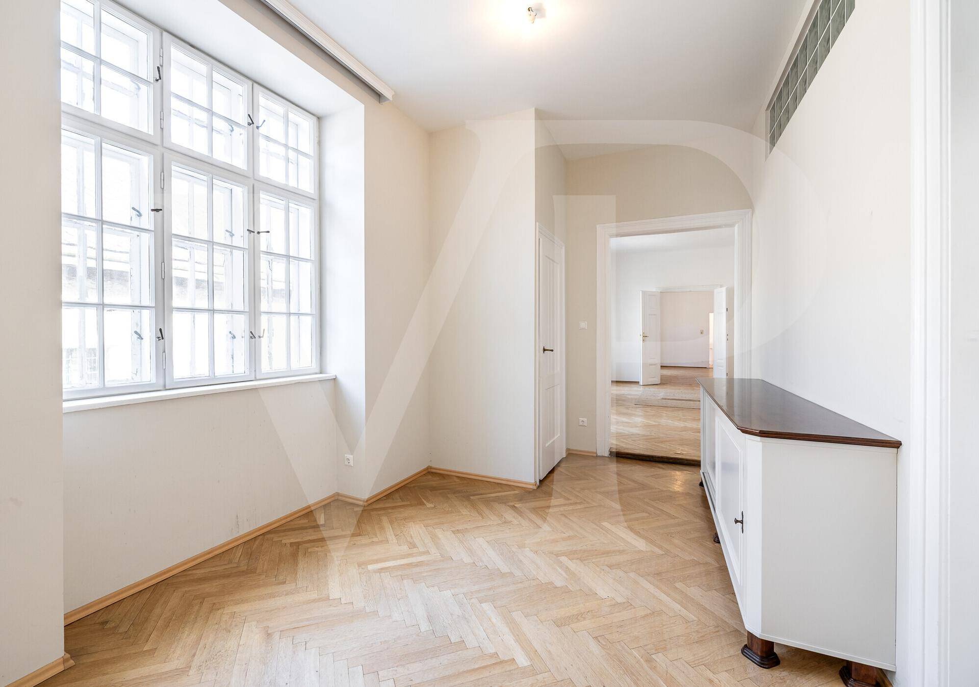 Helles Zimmer mit großen Fenstern, Fischgrätparkett und einem Durchgang zu weiteren Räumen.