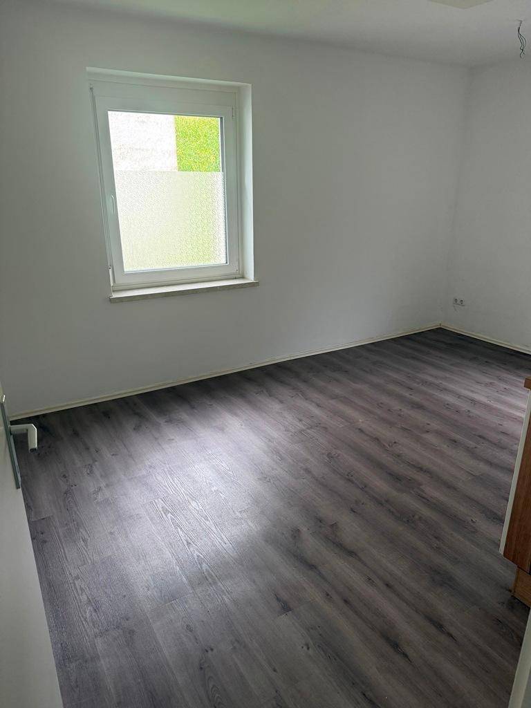 Leeres Zimmer mit Laminatboden, weißen Wänden und einem Fenster mit Blick ins Grüne.