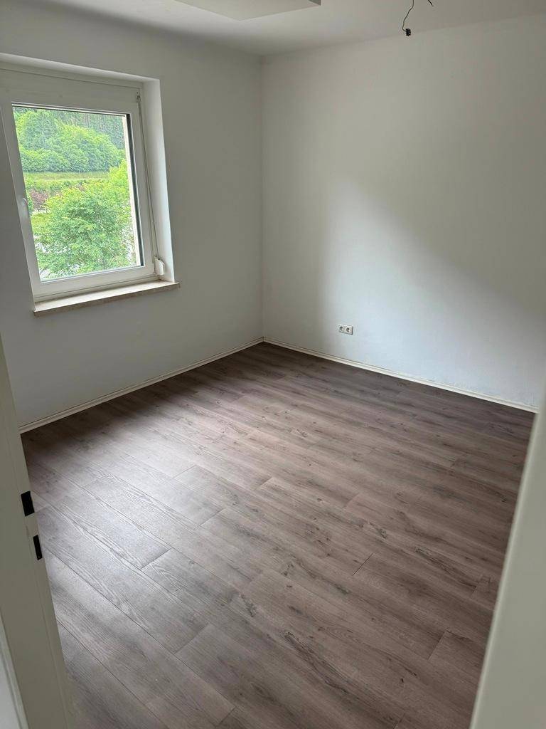 Helles Zimmer mit Laminatboden, weißen Wänden und einem großen Fenster mit grüner Aussicht.