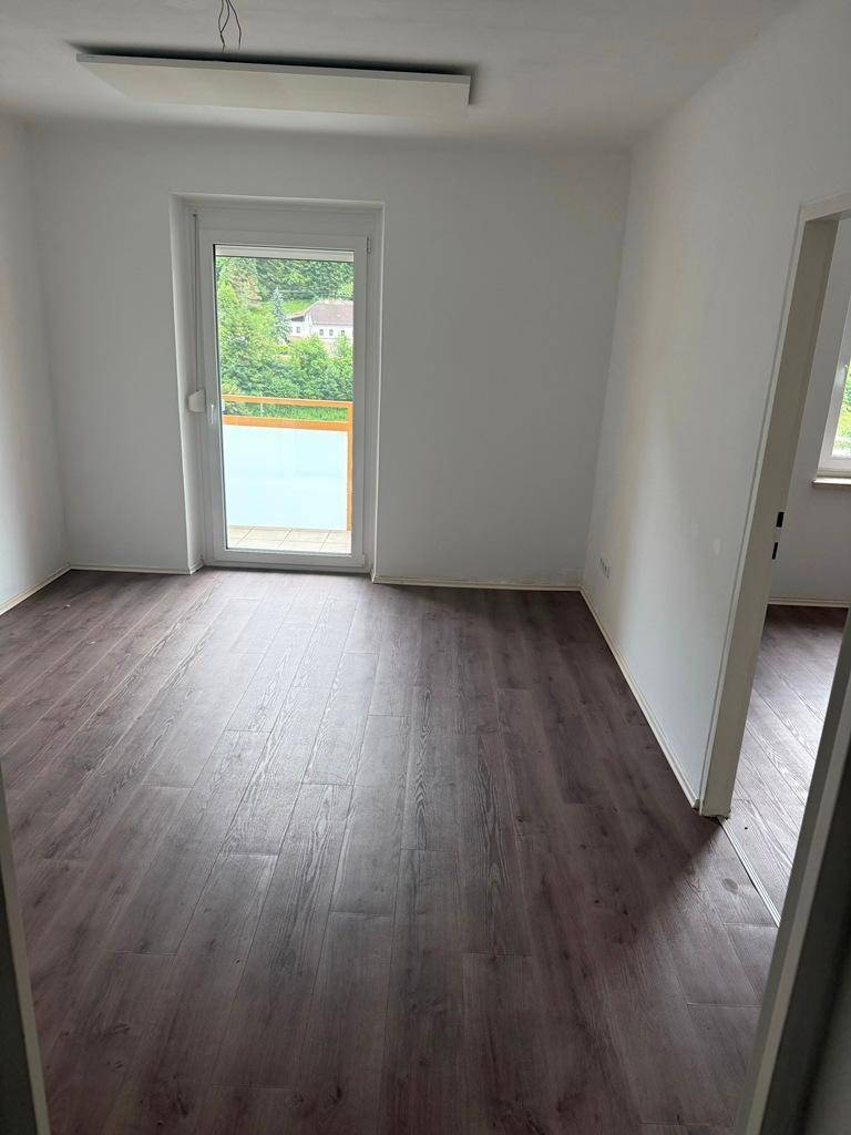 Heller Raum mit Laminatboden, weißen Wänden und Zugang zu einem Balkon mit grüner Aussicht.