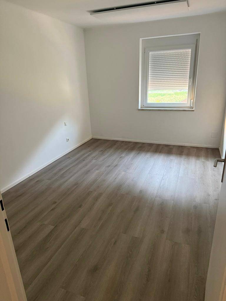 Leeres Zimmer mit hellem Laminatboden und einem Fenster mit Rollladen.