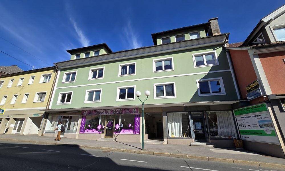 Mehrstöckiges Gebäude mit hellgrüner Fassade und Geschäften im Erdgeschoss an einer Straße.