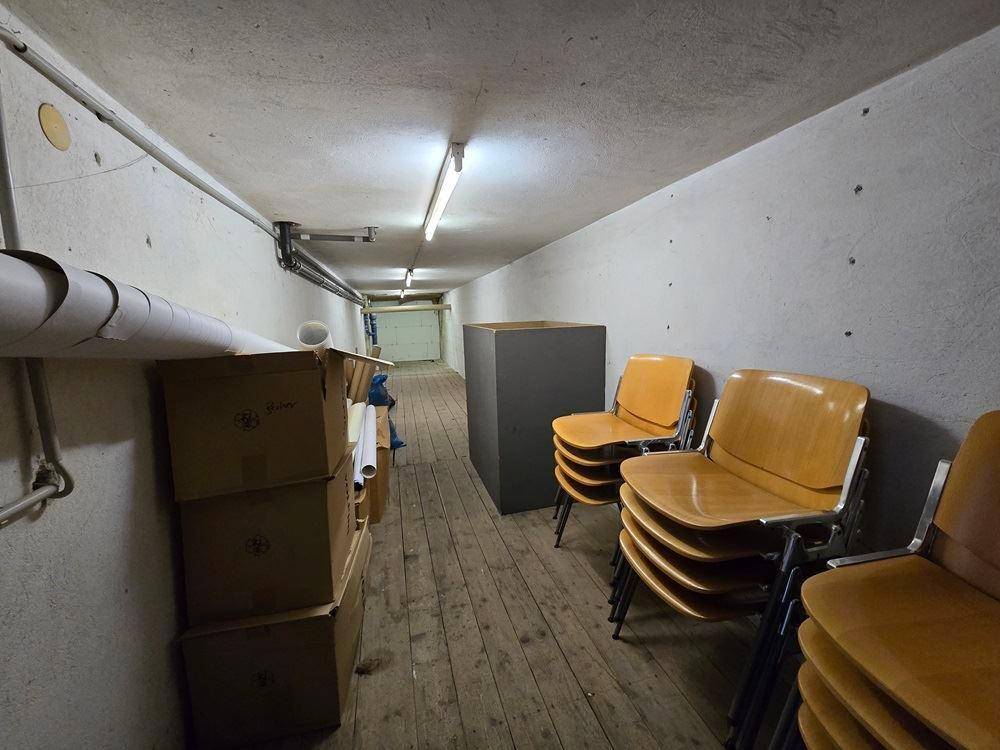 Langer Kellerraum mit Holzboden, niedriger Decke und gestapelten Stühlen und Kartons.