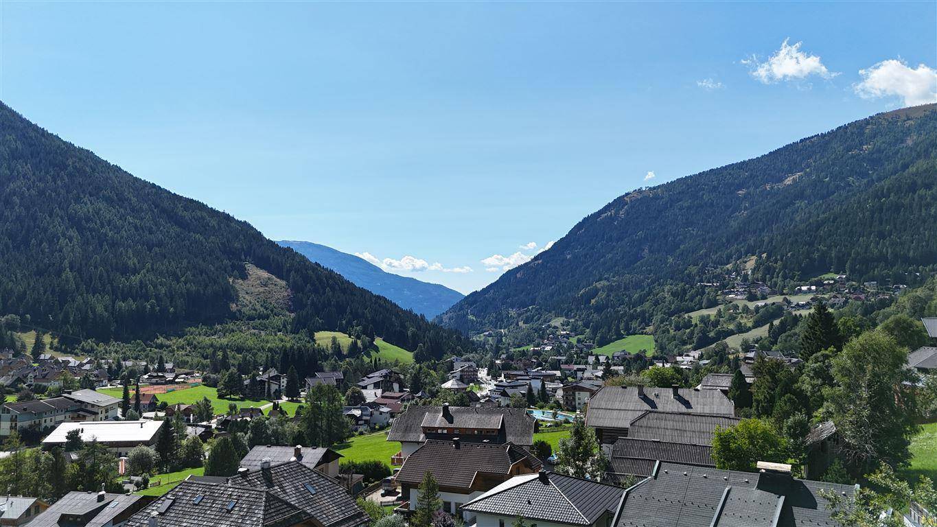 Weitläufiger Blick über ein Tal mit einem Dorf, eingebettet zwischen bewaldeten Bergen unter blauem Himmel.