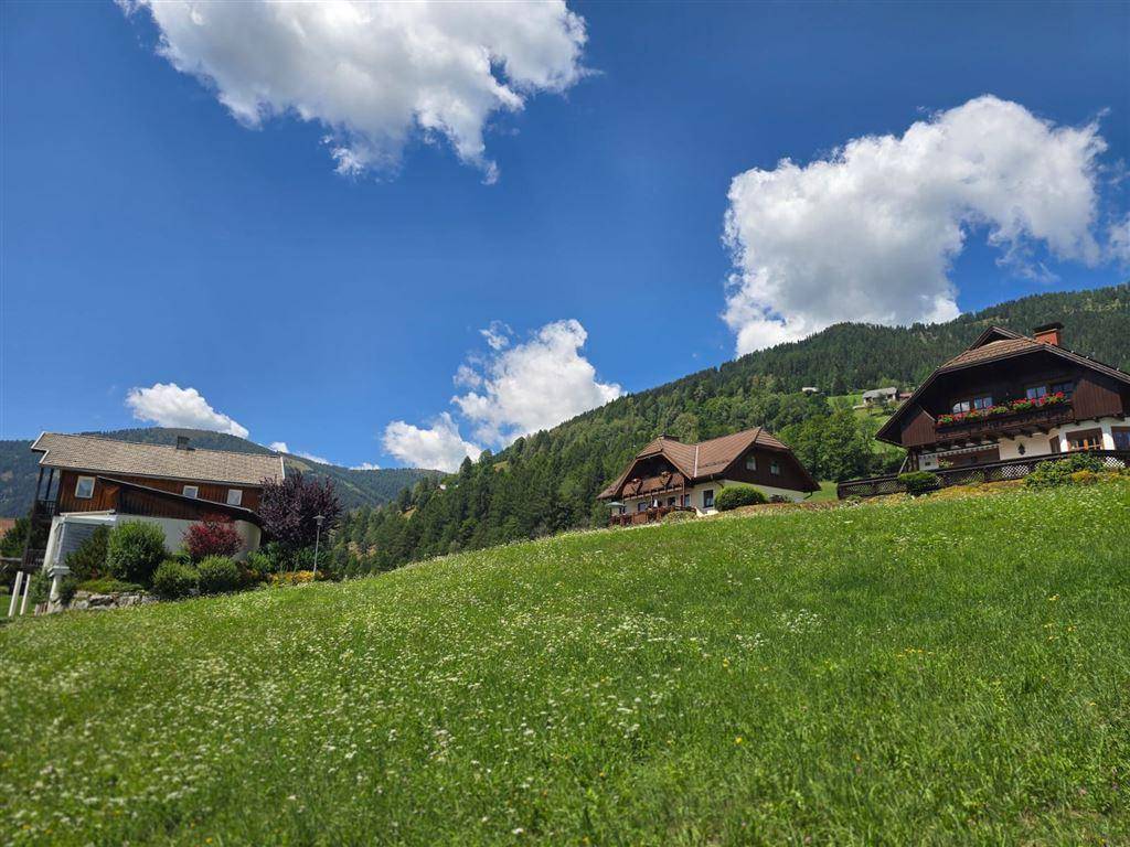 Häuser auf einem grünen Hügel mit weitem Blick auf die bewaldeten Berge unter blauem Himmel.