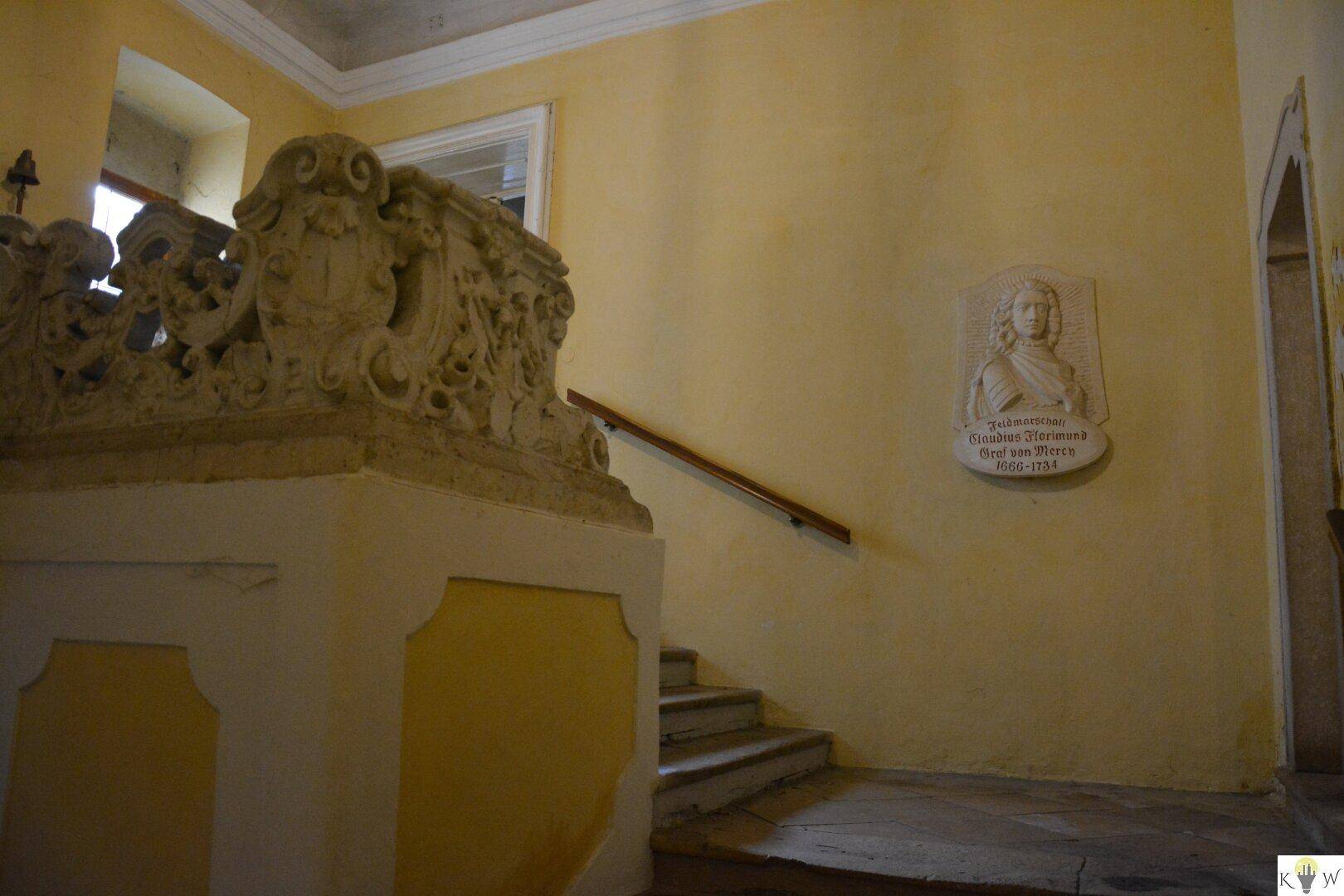 Historischer Treppenaufgang mit gelben Wänden, einer verzierten Steinbalustrade und einer Gedenktafel.