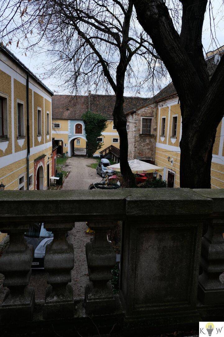 Blick von einer steinernen Balustrade auf einen historischen Innenhof mit gelben Gebäuden und Bäumen.