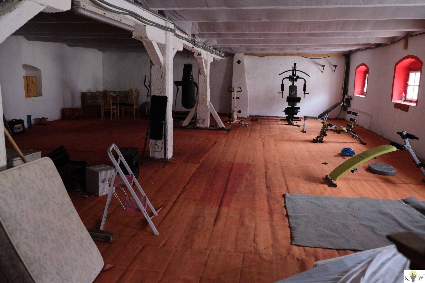 Großer Fitnessraum mit verschiedenen Trainingsgeräten, Holzbalken und roten Fenstern in rustikaler Umgebung.