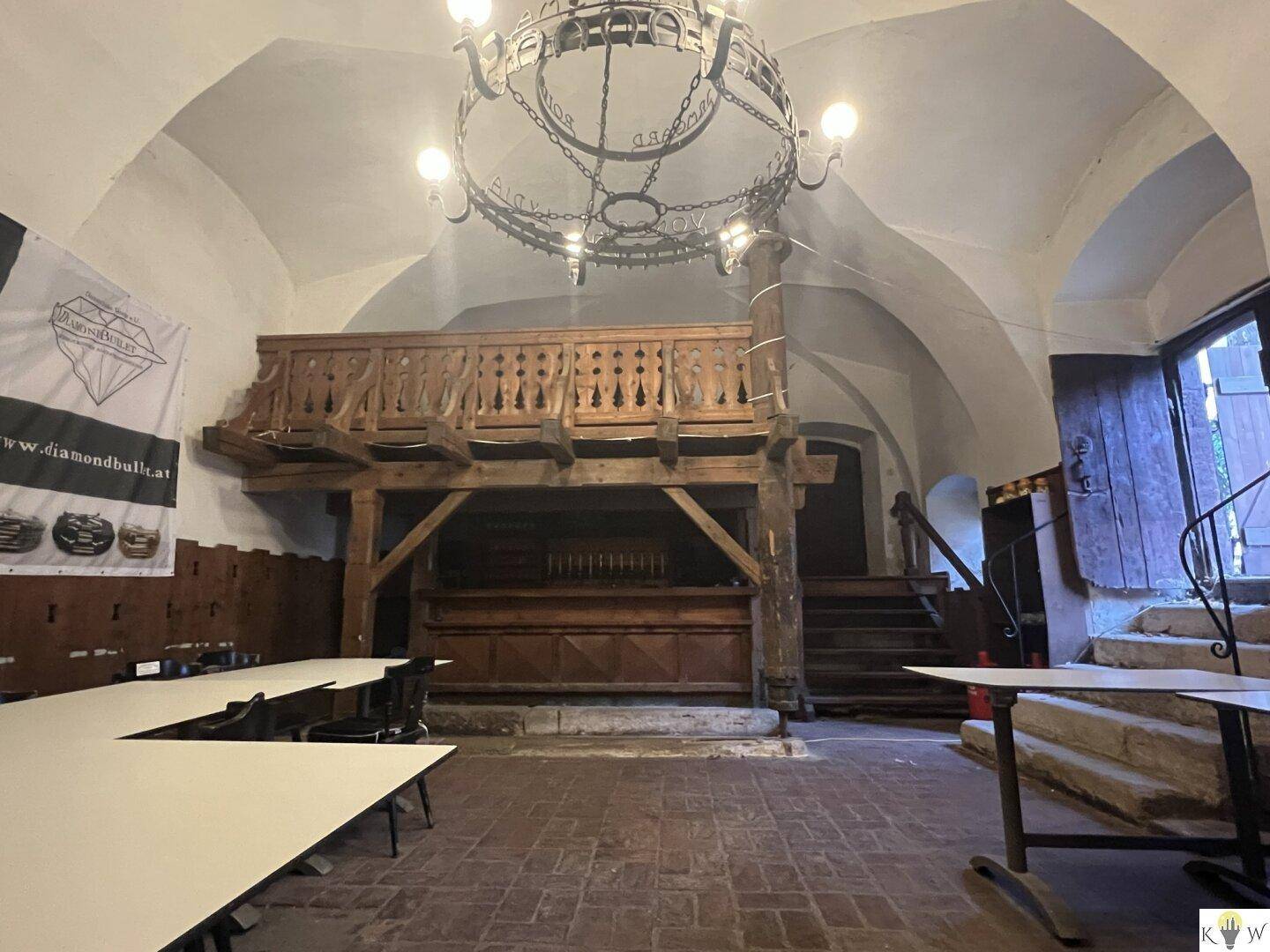 Großer Gewölberaum mit Holzbalkon, schmiedeeisernem Kronleuchter und Tischen für Veranstaltungen.