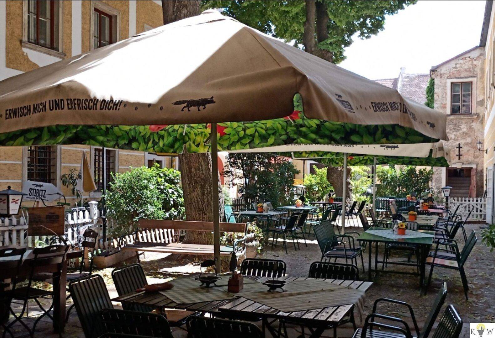 Sonniger Biergarten in einem Innenhof mit zahlreichen Tischen, Stühlen und großen Sonnenschirmen.