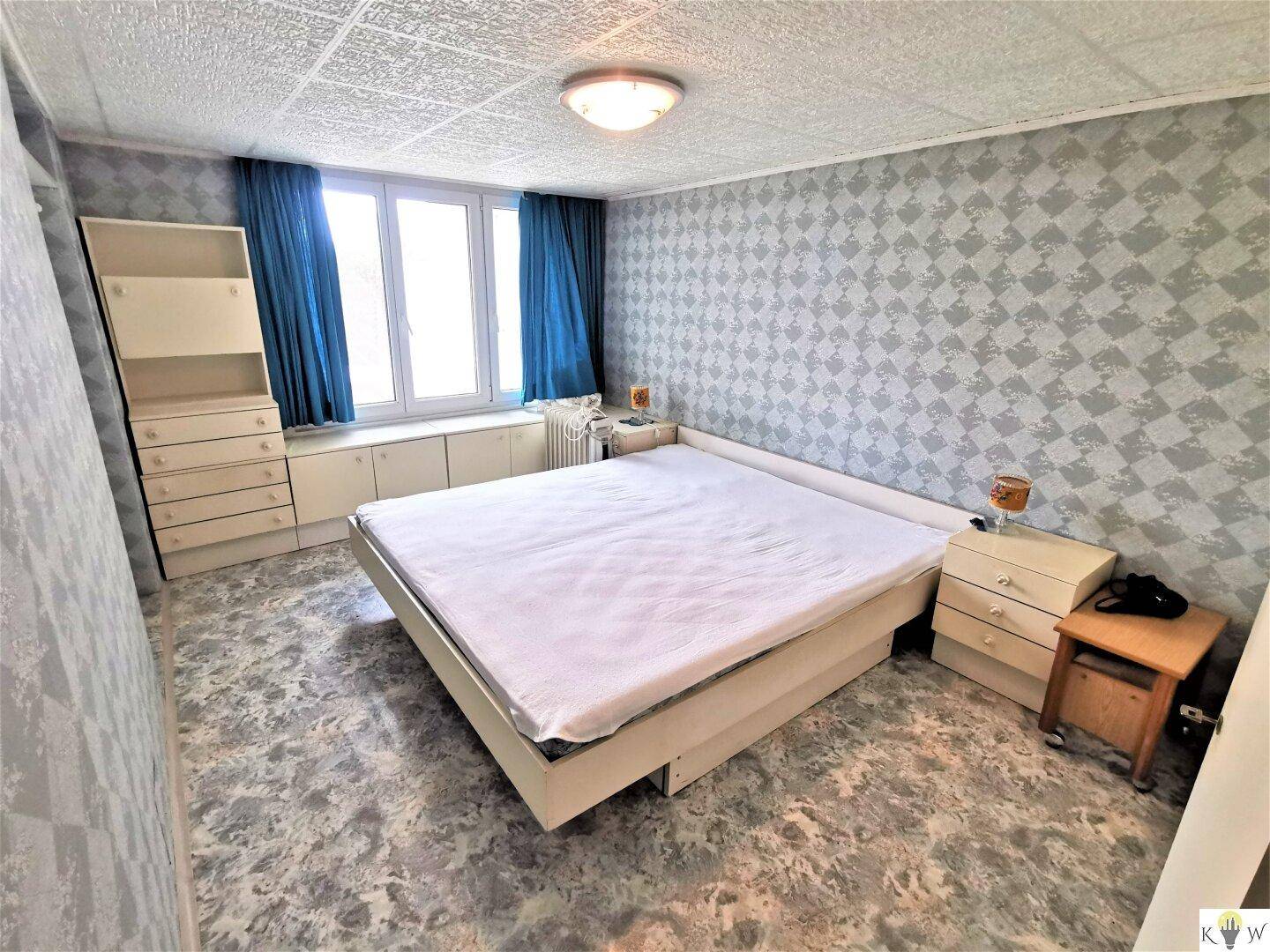 Schlafzimmer mit gemusterter Tapete, großem Bett und blauen Vorhängen am Fenster.