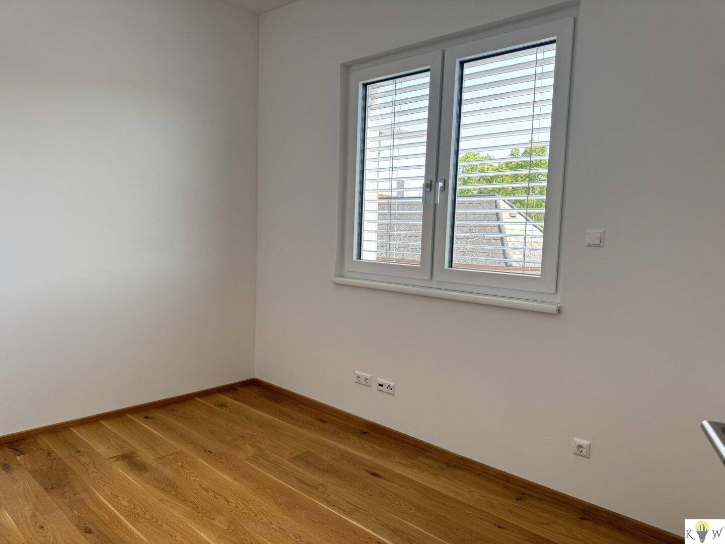Heller, leerer Raum mit Holzparkettboden, weißen Wänden und großem Fenster mit Jalousien.