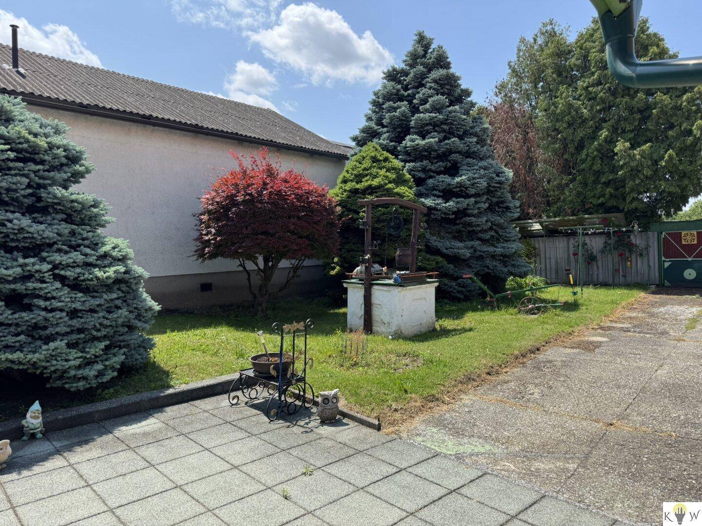 Gepflasterter Außenbereich mit Gartenfiguren, Nadelbäumen und einem dekorativen Brunnen.