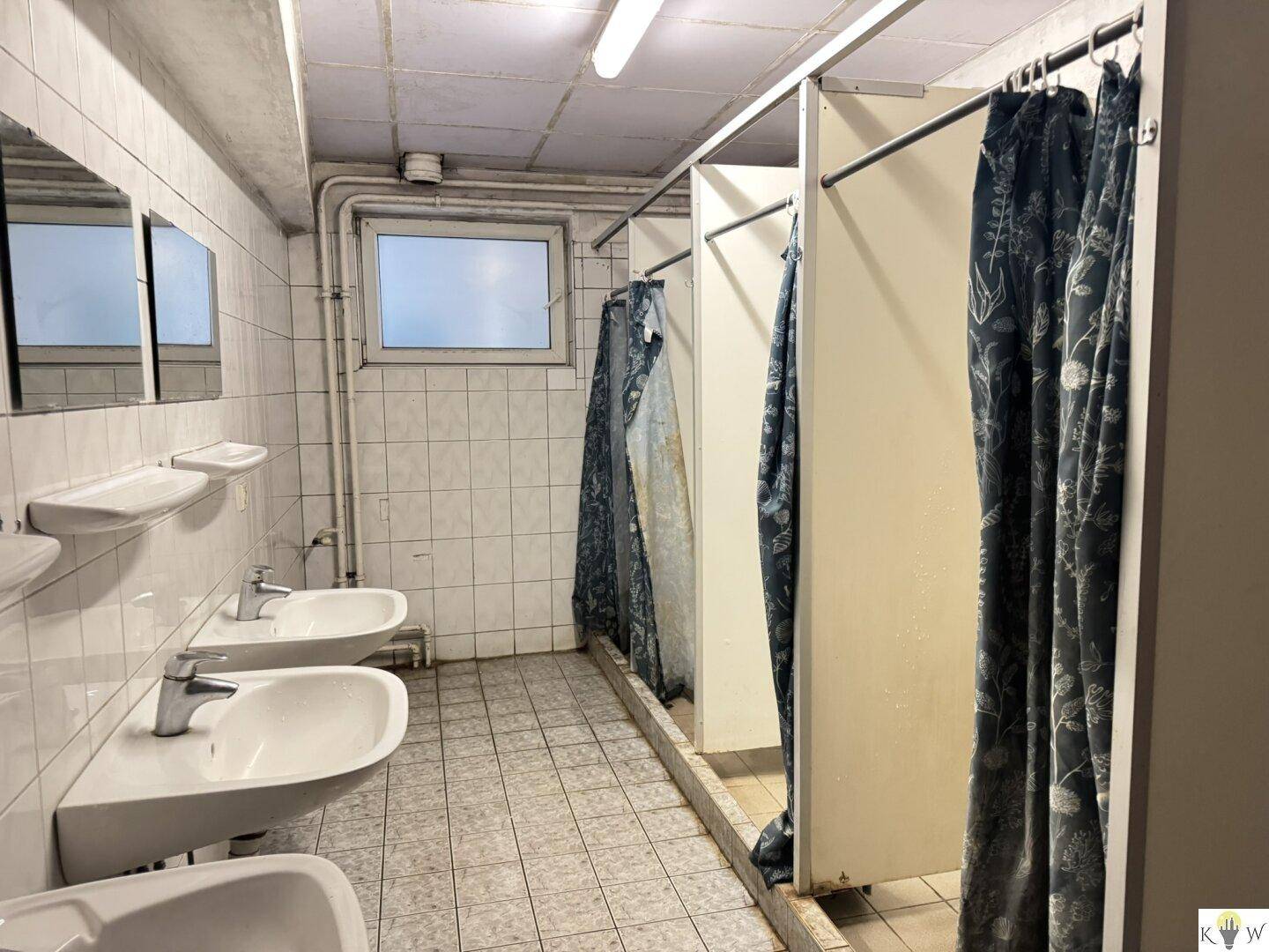 Waschraum mit mehreren Waschbecken und Duschkabinen, beleuchtet durch ein kleines Fenster.
