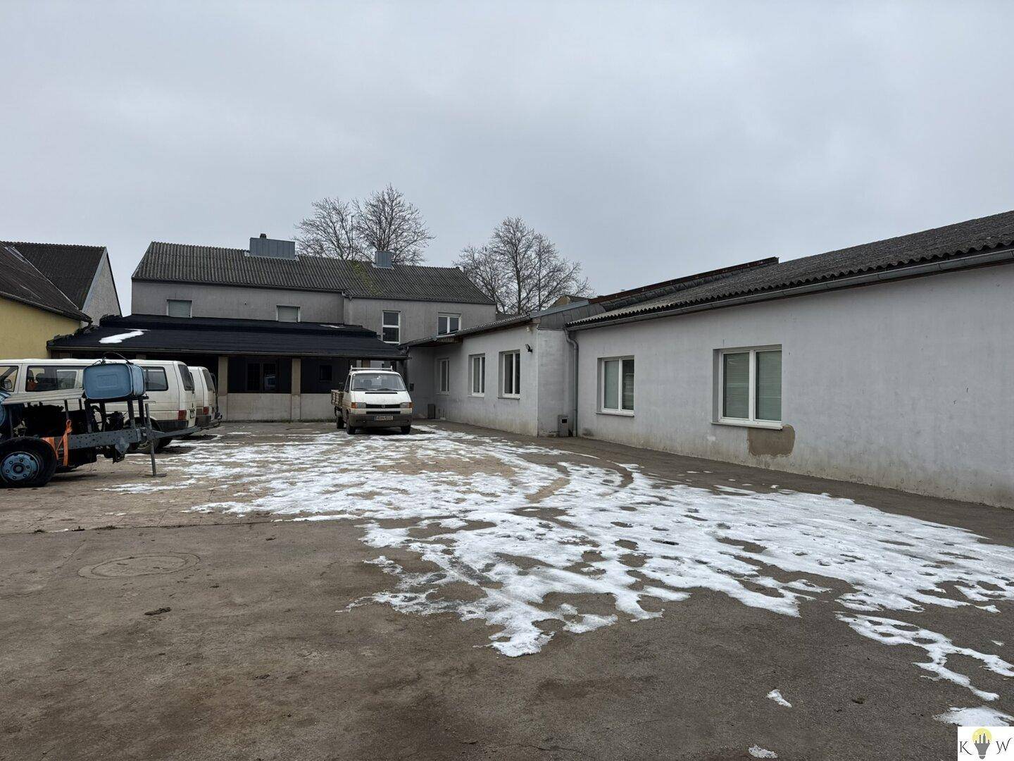 Weitläufiger Außenbereich mit mehreren Gebäuden, geparkten Fahrzeugen und Schneeresten auf dem Asphalt.