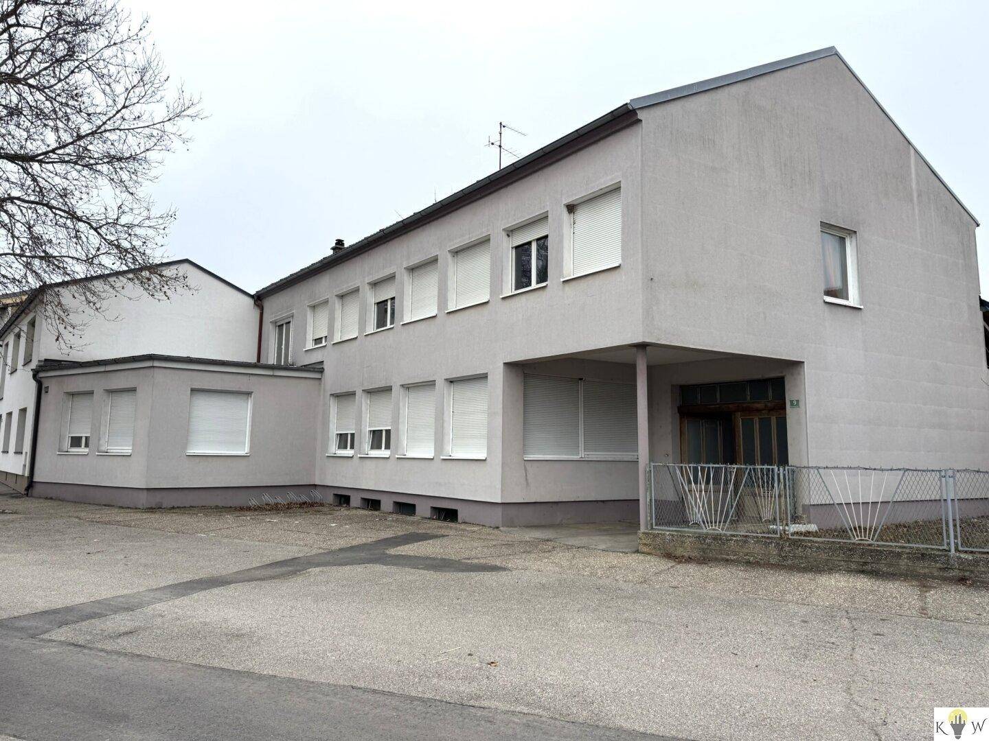 Ansicht des grauen Gebäudes mit mehreren Fenstern und Rollläden, einem kleinen Zaun und einem Vorplatz.