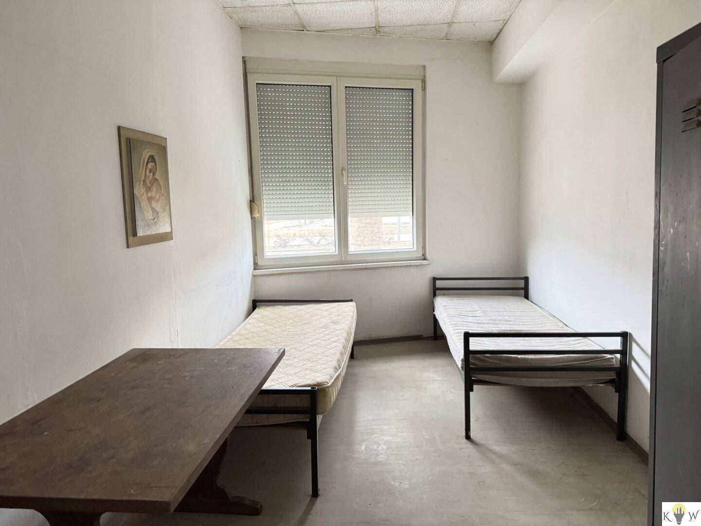 Schlichtes Schlafzimmer mit zwei Einzelbetten, einem Holztisch und einem Fenster mit Blick nach draußen.