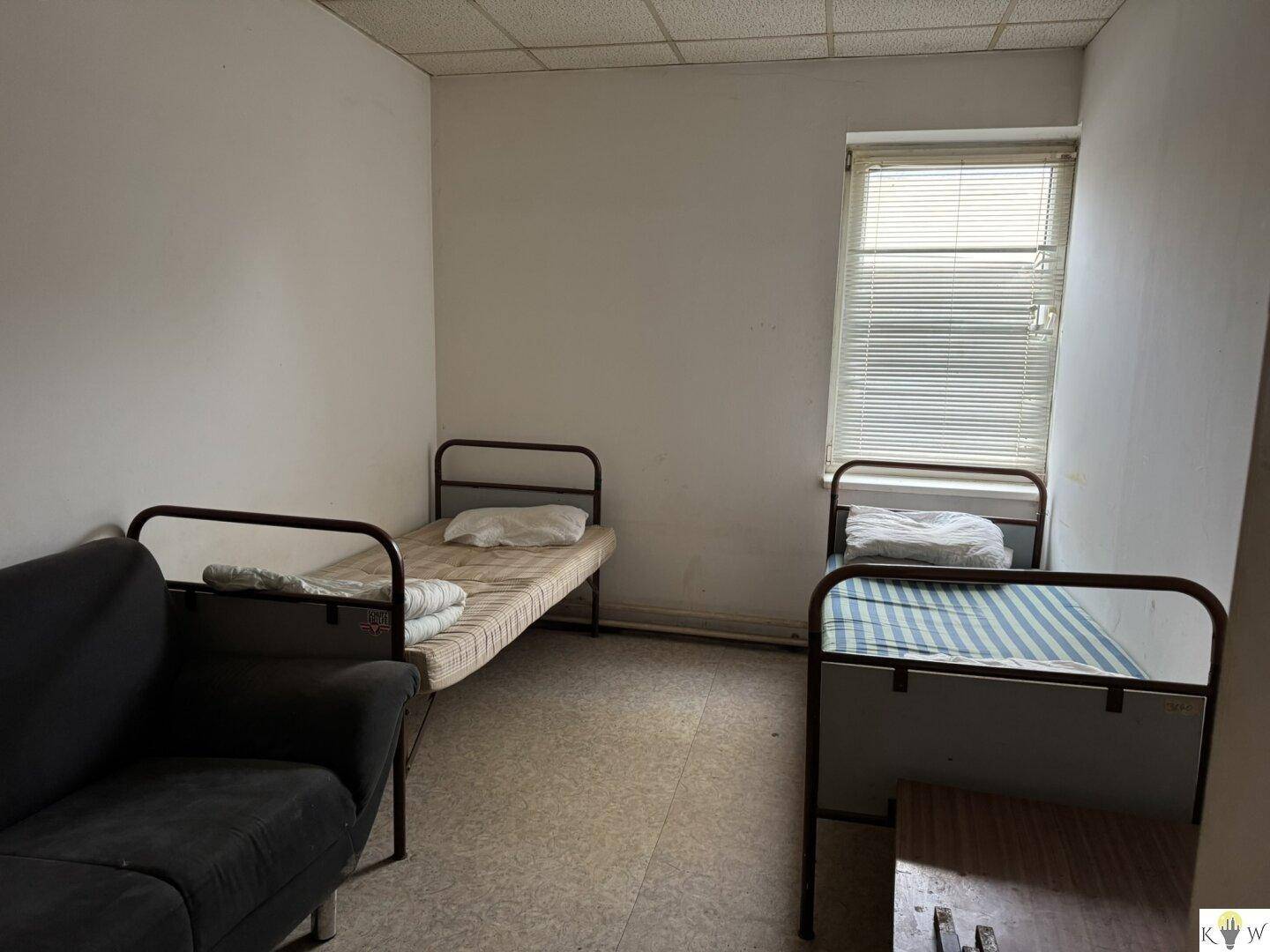 Einfaches Schlafzimmer mit zwei Einzelbetten und einem Sofa, beleuchtet durch ein Fenster mit Jalousien.