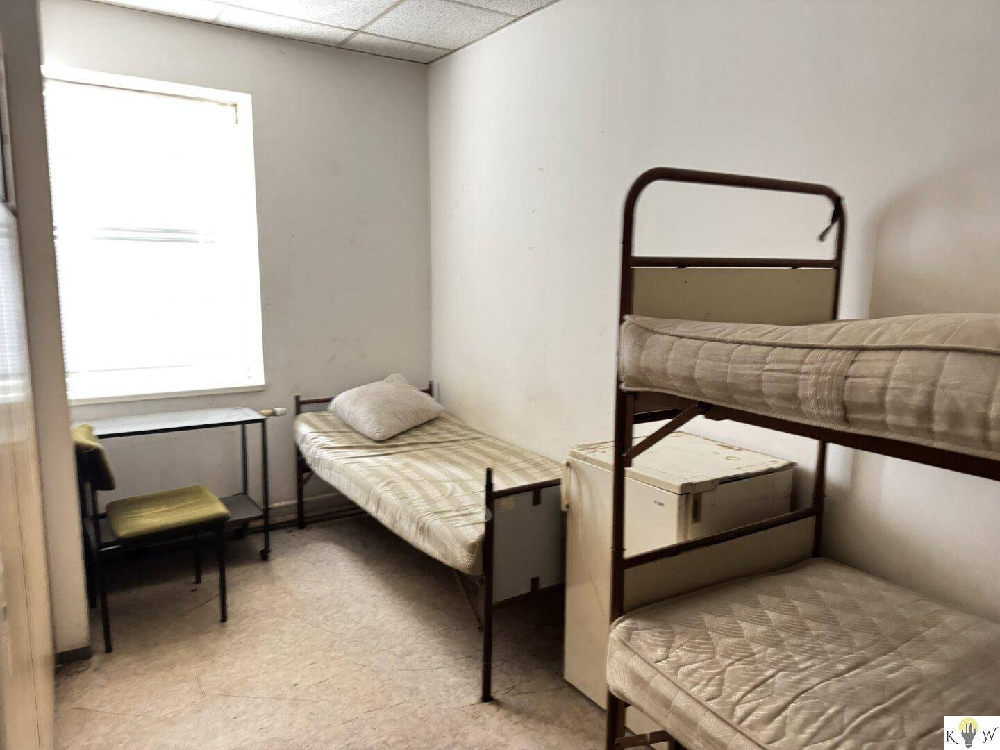 Schlichtes Schlafzimmer mit einem Einzelbett und einem Etagenbett, ergänzt durch einen kleinen Kühlschrank und ein Fenster.