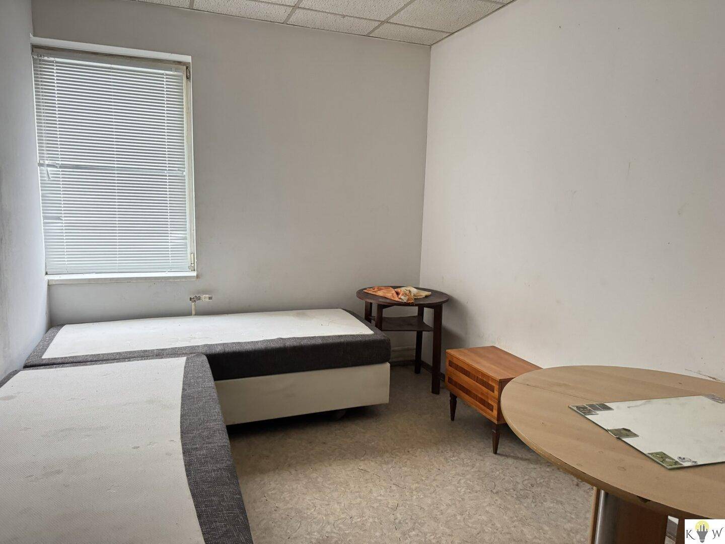 Schlichtes Schlafzimmer mit zwei Einzelbetten und einem runden Tisch, beleuchtet durch ein Fenster mit Jalousien.