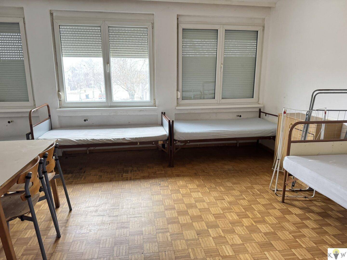 Geräumiges Schlafzimmer mit mehreren Betten und einem Parkettboden, beleuchtet durch große Fenster.