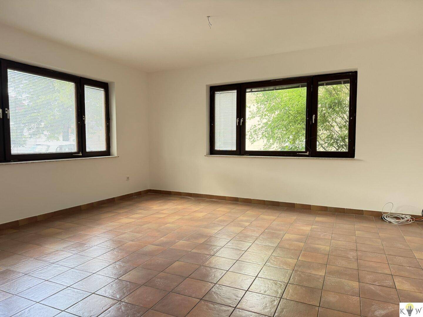 Geräumiges Zimmer mit Terrakottafliesen und zwei großen Fenstern mit Blick ins Grüne.