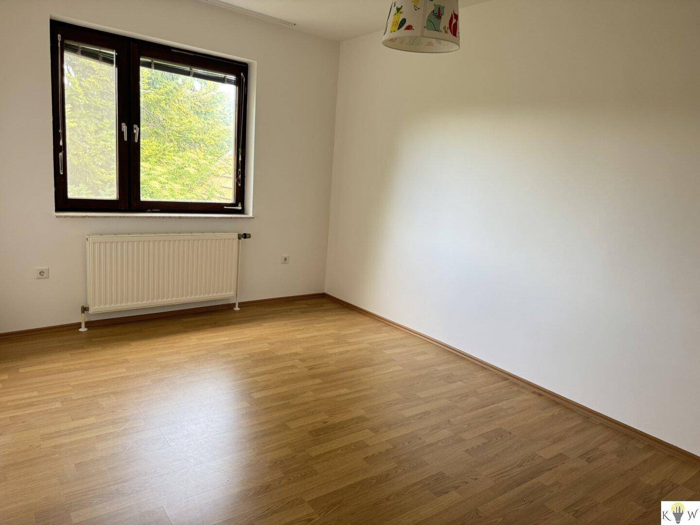 Helles Schlafzimmer mit Laminatboden, Heizkörper und Fenster mit Blick ins Grüne.