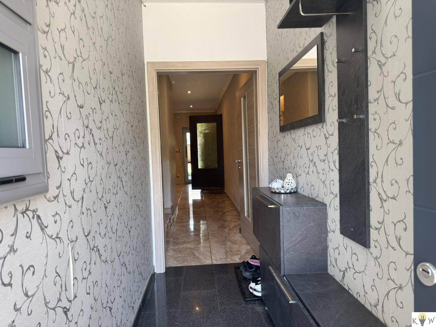 Heller Flur mit gemusterter Tapete, dunklem Fliesenboden und einer Garderobe mit Spiegel.